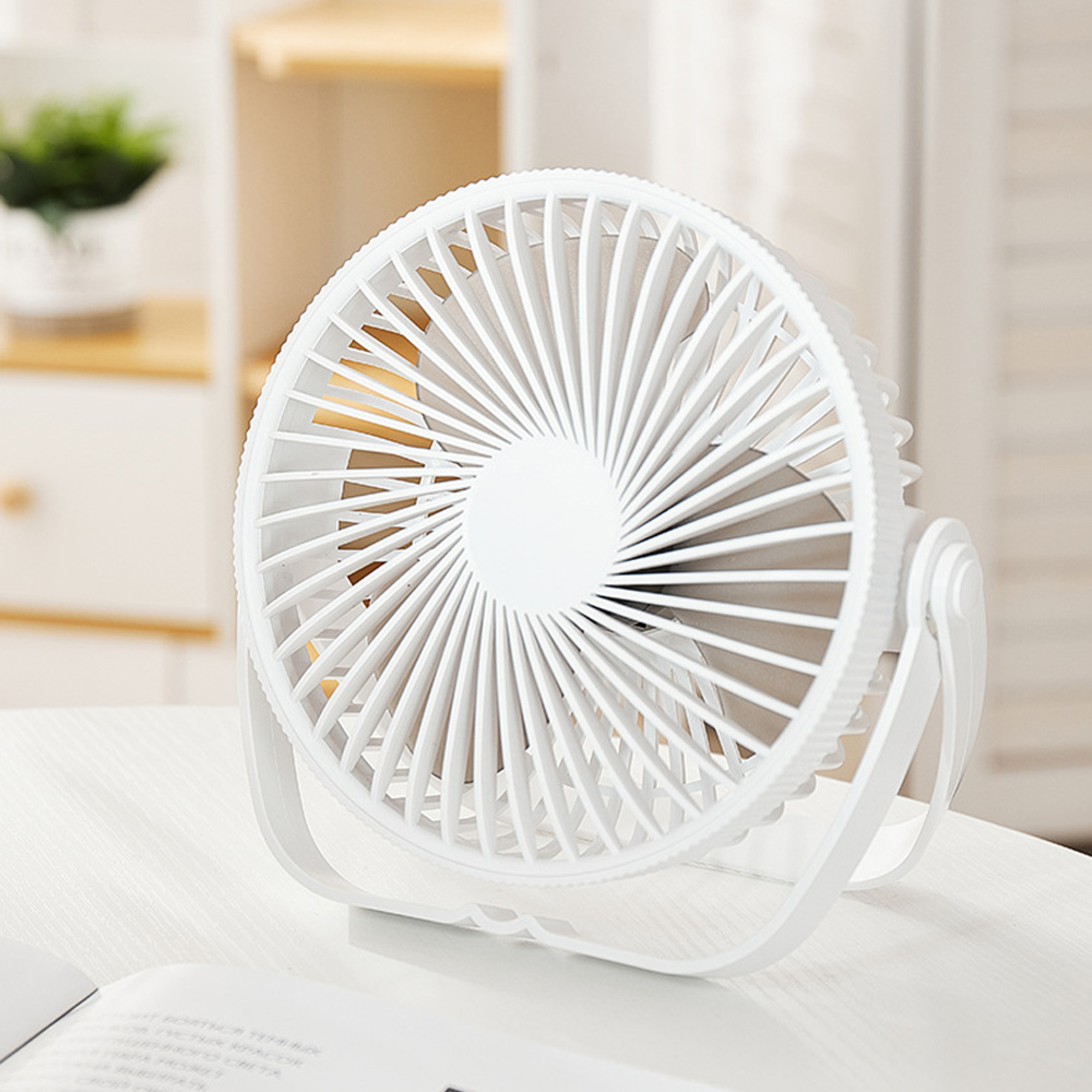 Ein weißer kleiner Ventilator steht auf einem Tisch. Im Hintergrund ist ein Regal mit einer Pflanze.