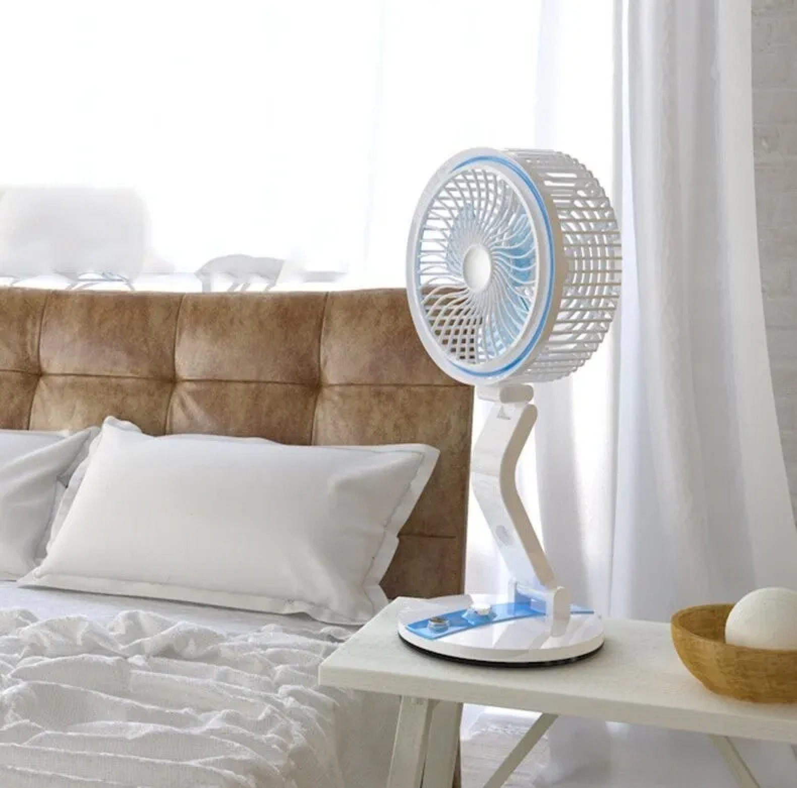 Ein weißer Ventilator mit blauem Rand steht auf einem Nachttisch neben einem Bett.