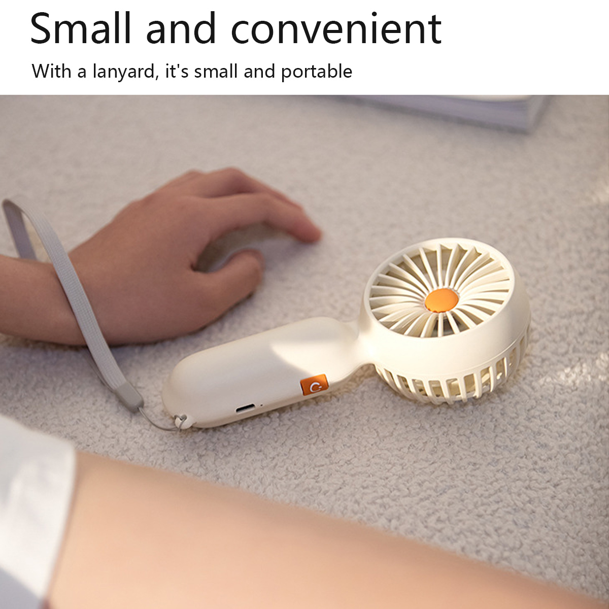 Ein weißer tragbarer Ventilator mit orangefarbenem Mittelteil liegt auf einer strukturierten Oberfläche, gehalten von einer Hand mit Lanyard.