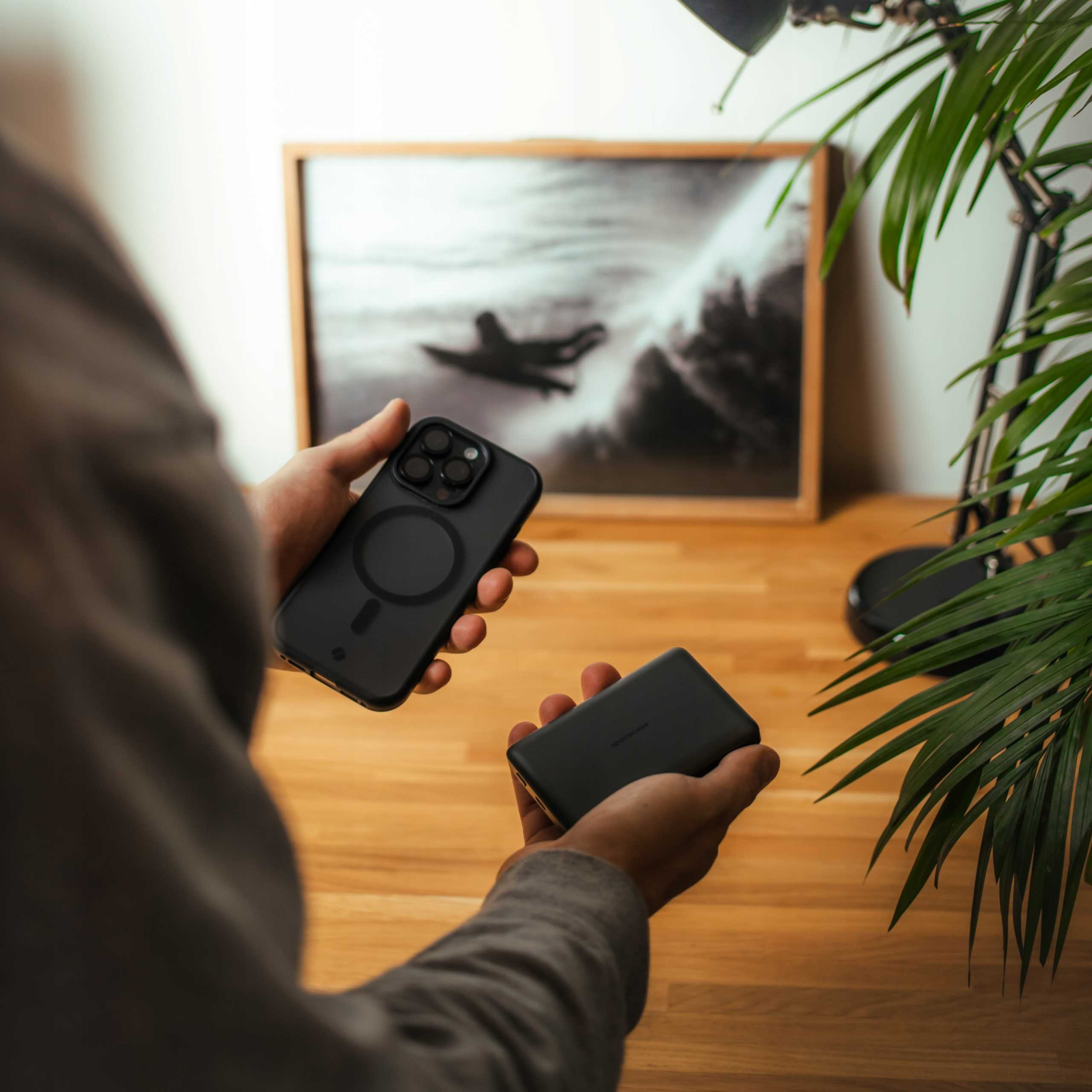 Osoba trzyma czarną smartfon i powerbank na drewnianym stole.