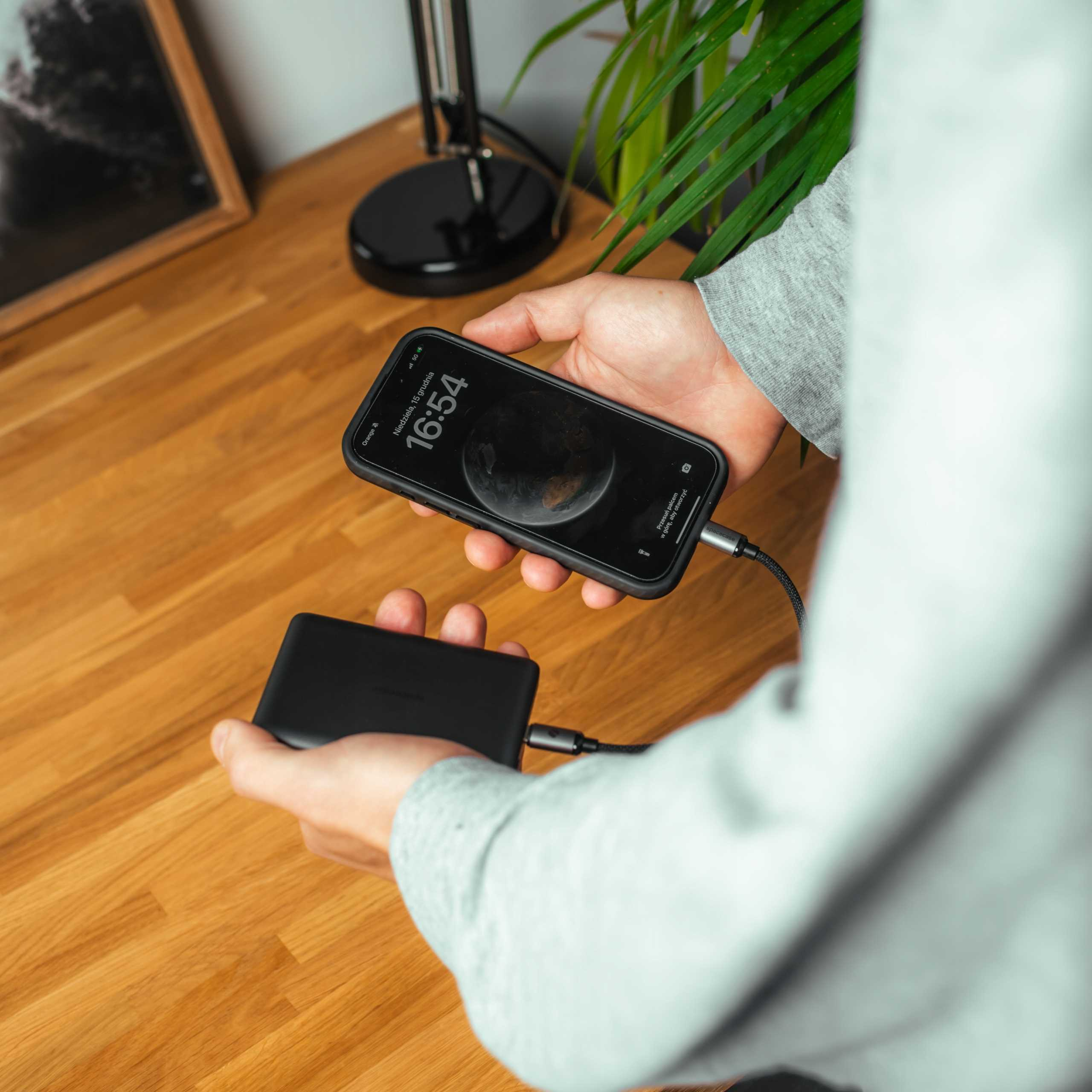 Osoba ładuje smartfon czarnym powerbankiem na drewnianym biurku.