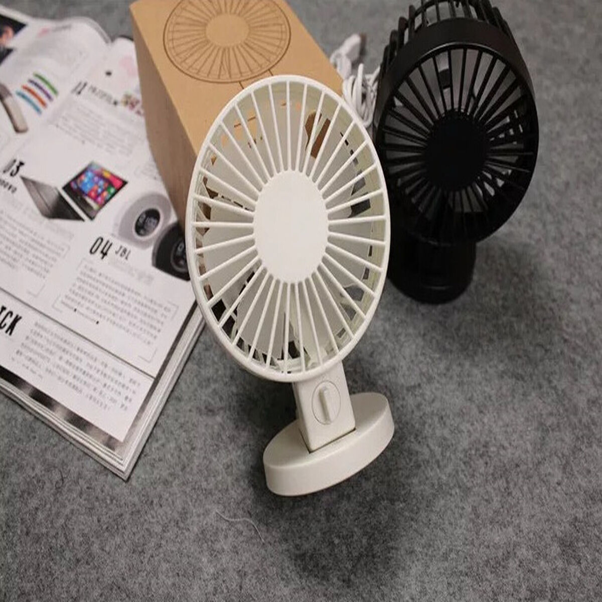 Zwei tragbare Ventilatoren, einer weiß und einer schwarz, stehen auf grauer Oberfläche neben einer offenen Zeitschrift.
