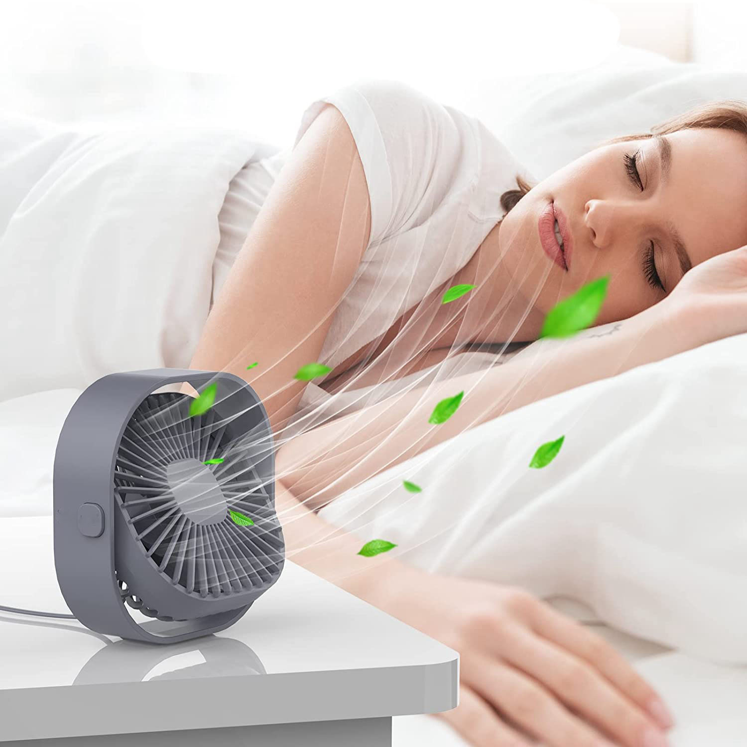 Ein grauer Schreibtischventilator bläst Luft mit grünen Blättern auf eine schlafende Frau im Bett.