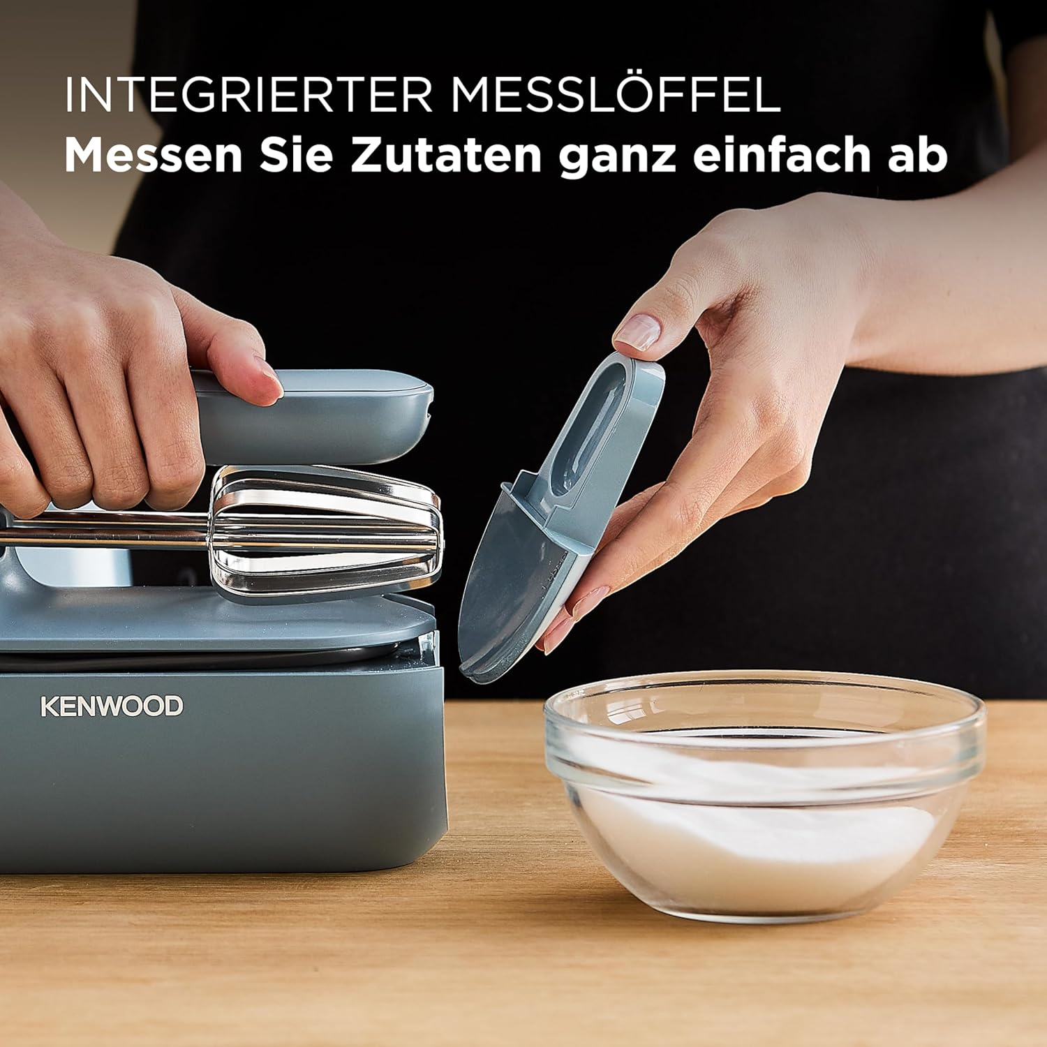Eine Hand hält einen grauen Messlöffel über einer Küchenmaschine mit Rührbesen.
