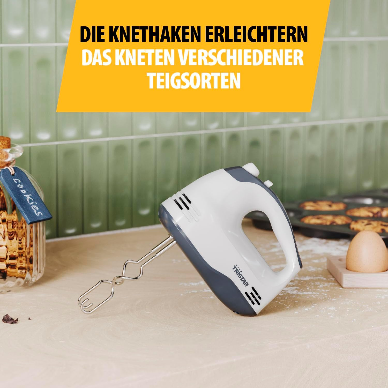 Ein Handmixer aus Weiß und Grau liegt auf einem Holztisch neben einem Glas Keksen.