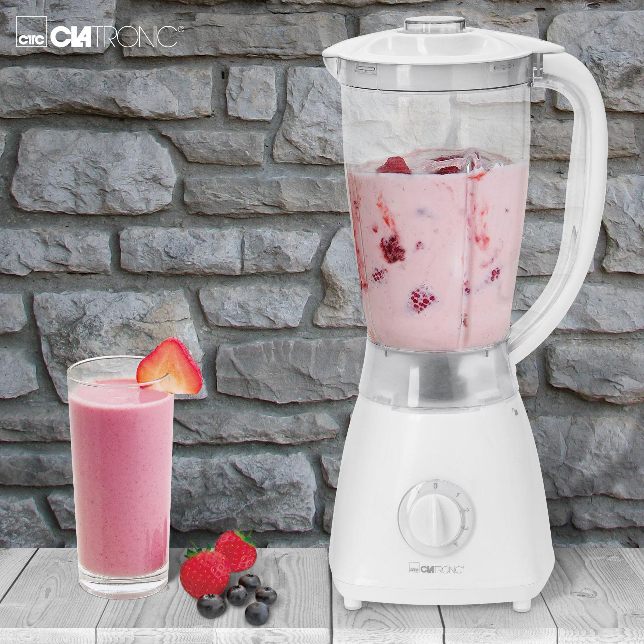 Ein weißer Mixer mit einem Glas Erdbeer-Smoothie und Früchten daneben vor einer Steinwand.
