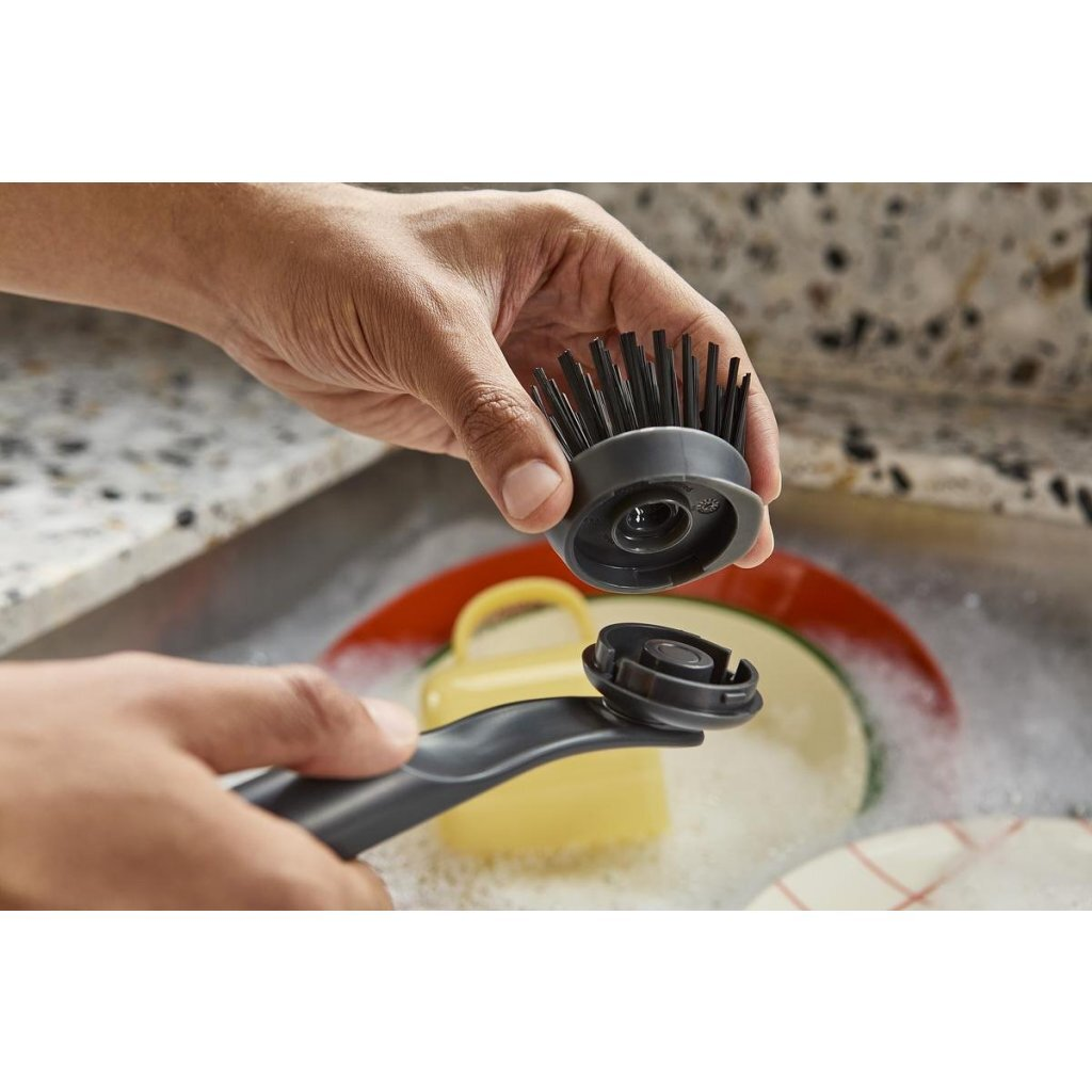 Manos ensamblan un cabezal de cepillo para platos gris en un mango sobre un fregadero con platos.