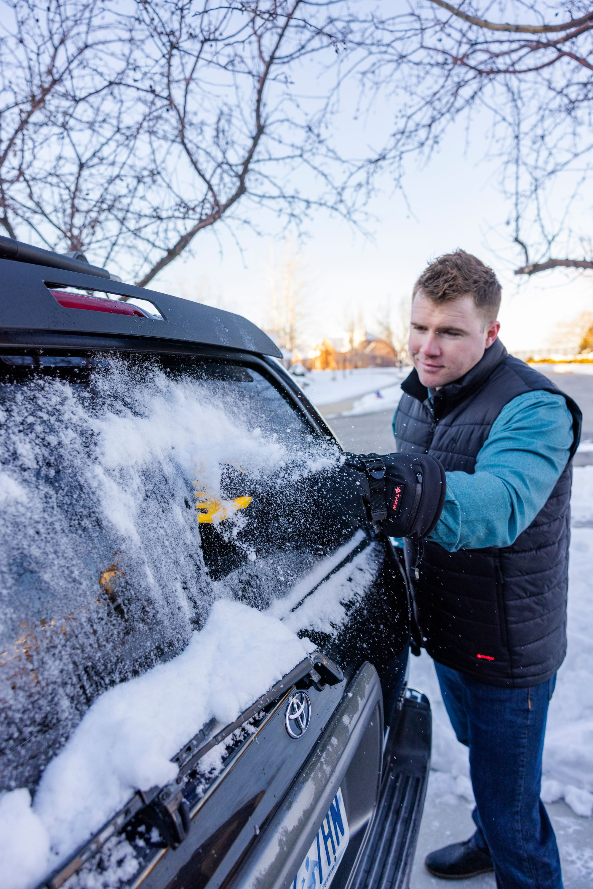 Un uomo in abiti invernali raschia la neve dal lunotto posteriore di un'auto.