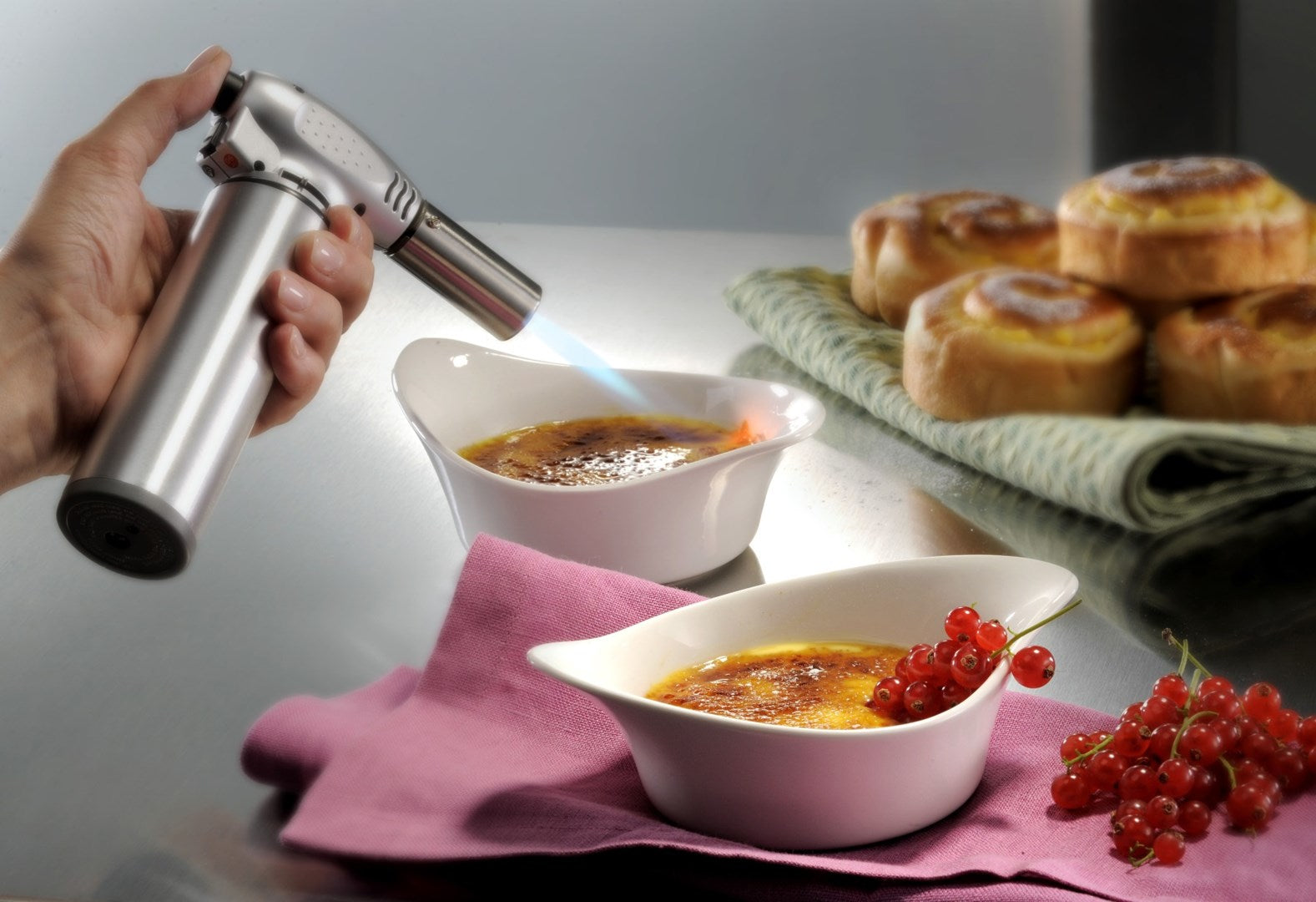 Eine Hand benutzt einen Brenner, um Creme Brulee zu karamellisieren. Daneben sind Gebäck und rote Johannisbeeren.