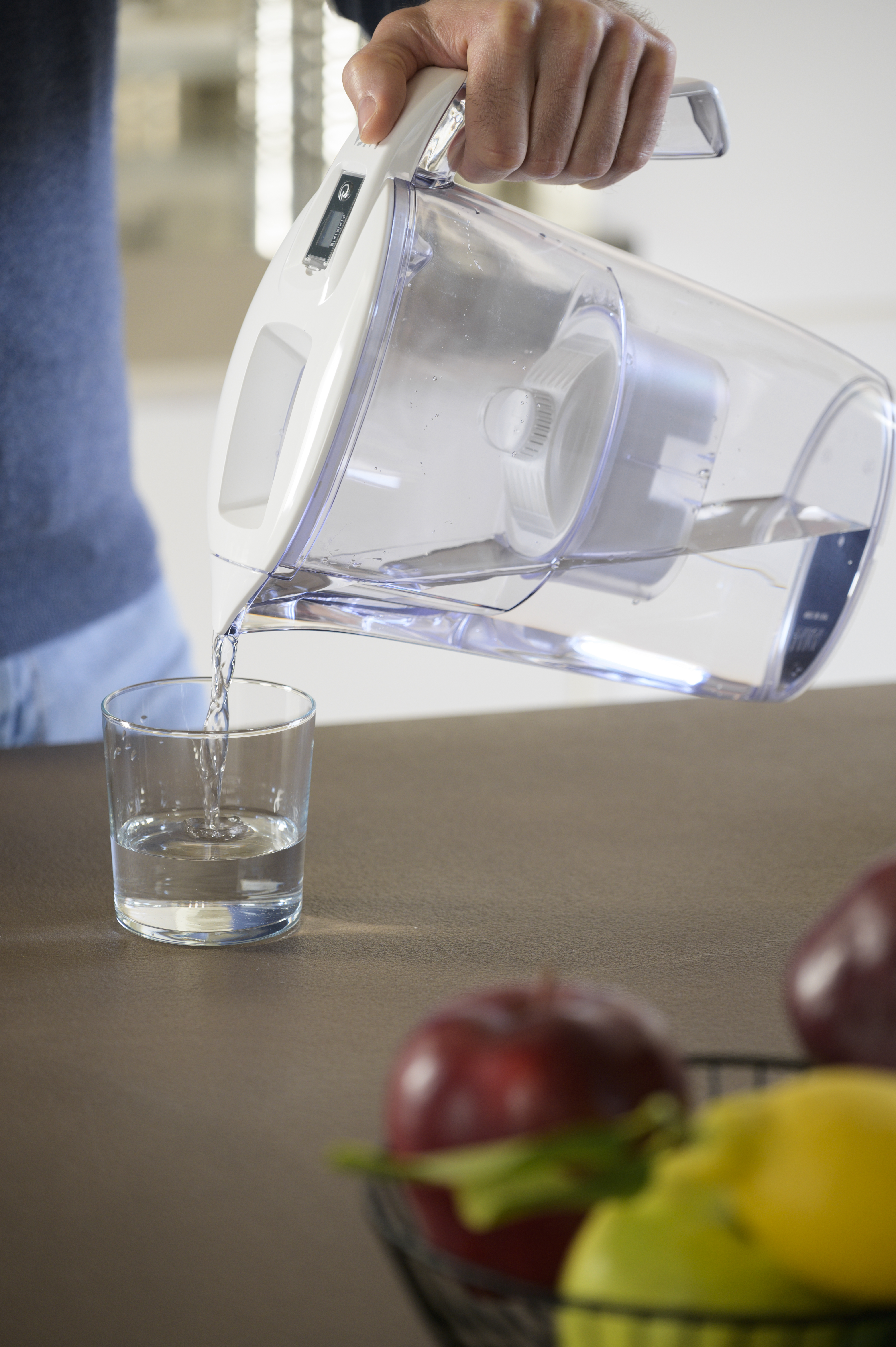 Una mano vierte agua de una jarra con filtro en un vaso. Hay fruta delante.