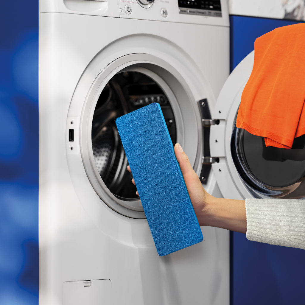 Mano che tiene mattone pulente blu vicino a lavatrice aperta con maglia arancione all'interno.