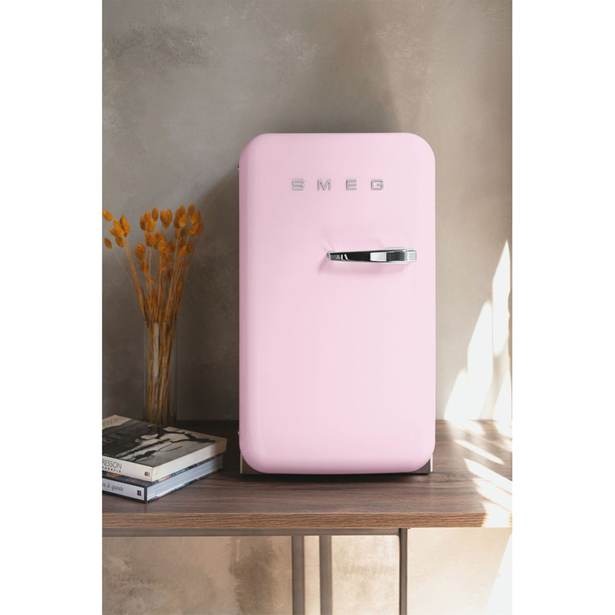 Ein kleiner, pastellrosa Smeg-Kühlschrank steht auf einem Holztisch neben Büchern und getrockneten Blumen.