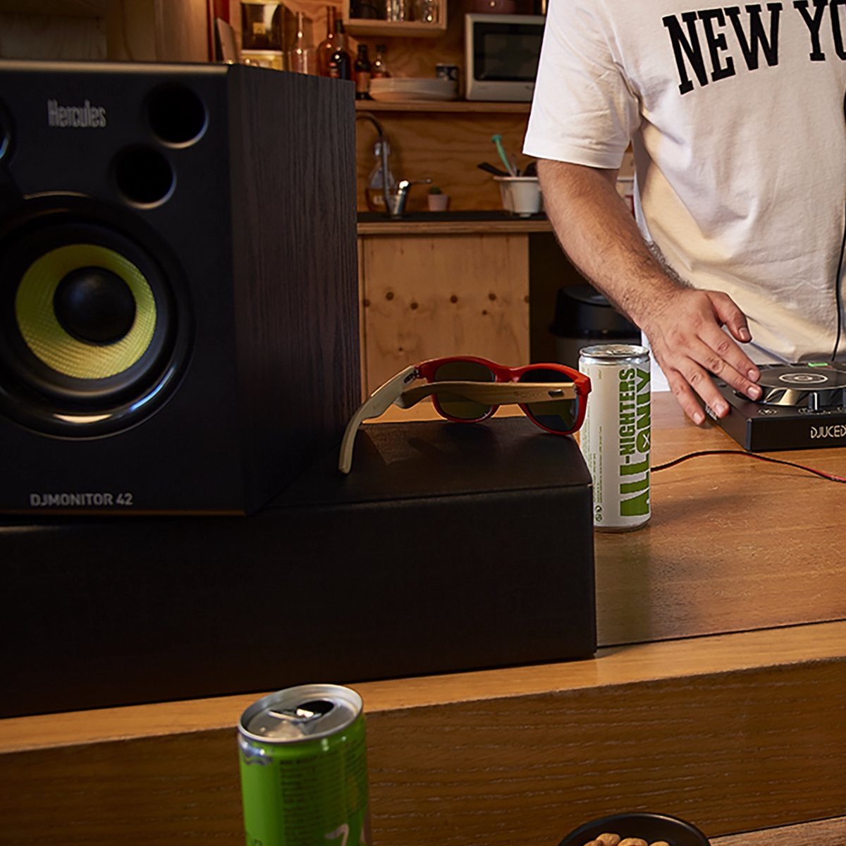 Ein DJ-Monitorlautsprecher, rote Sonnenbrille und Getränkedosen auf einer Holztheke.