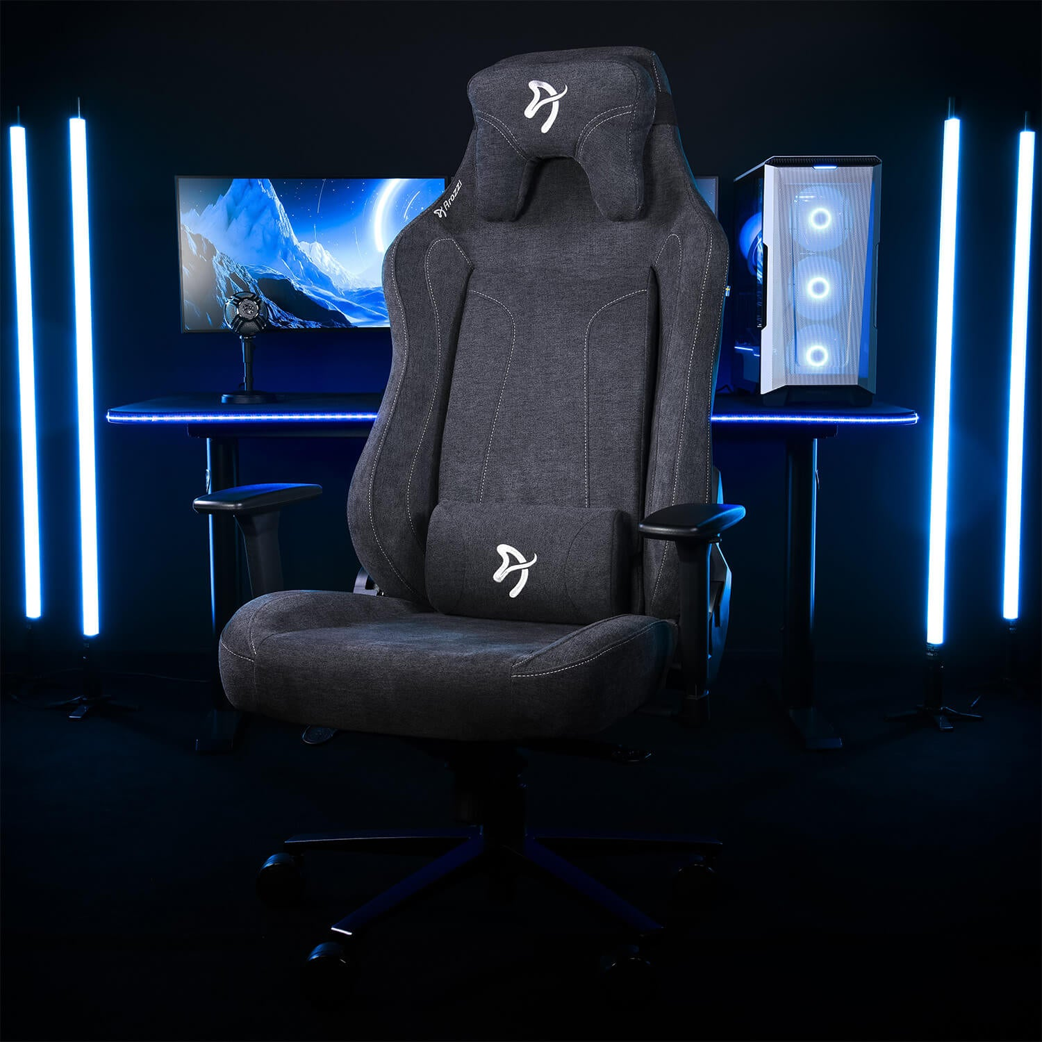 Een donkere gamestoel staat voor een bureau met computer en monitor. Blauwe lichten zijn aanwezig.