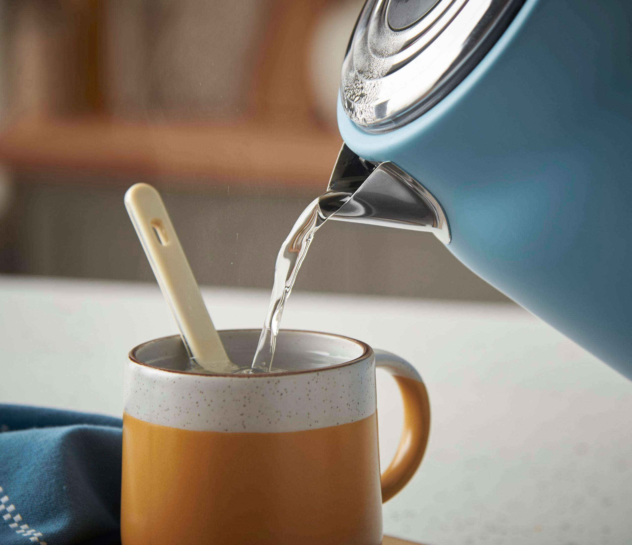 Blauer Wasserkocher gießt heißes Wasser in eine gelbe Tasse mit Löffel.
