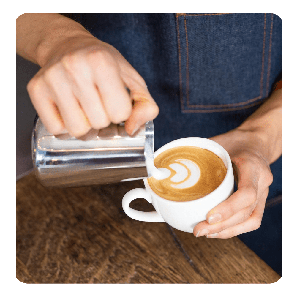 Barista gießt Latte Art in eine weiße Tasse. Ein Milchkännchen wird benutzt.