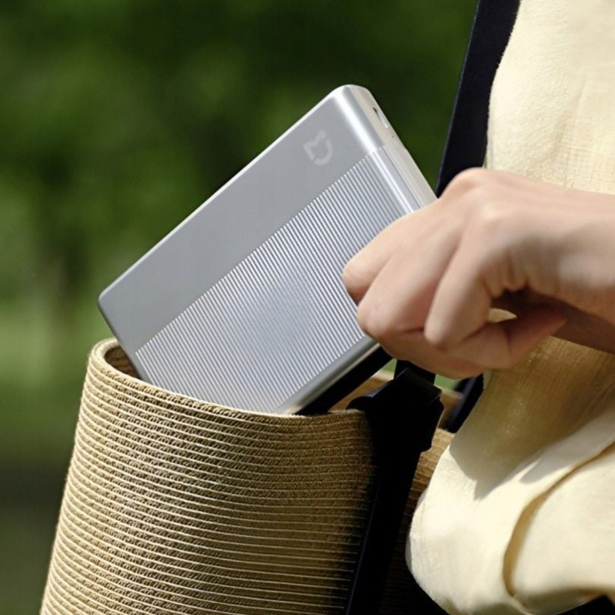 Una mano inserisce un power bank argentato in un cappello di paglia. Sfondo verde sfocato.
