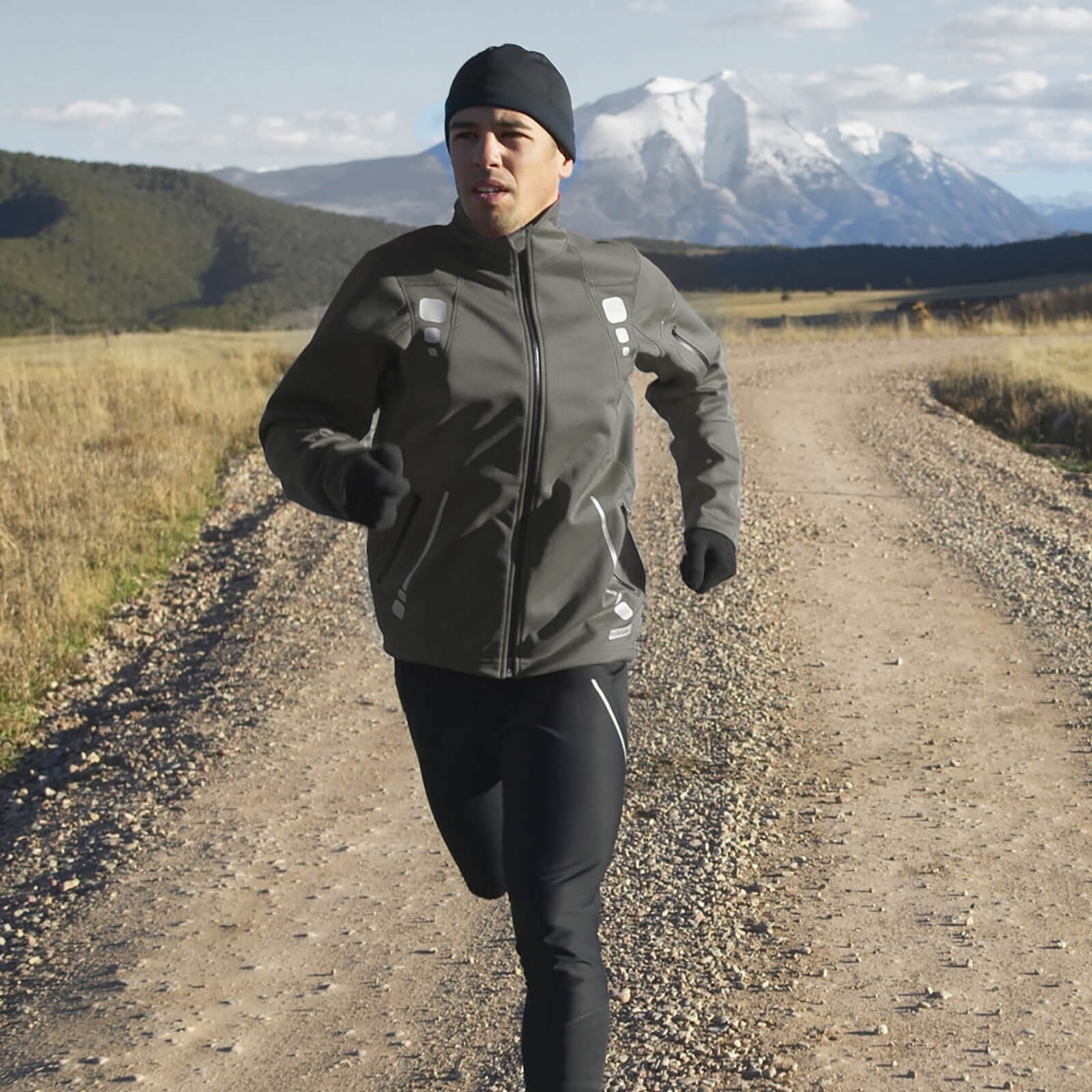 Un uomo in abbigliamento sportivo corre su un sentiero sterrato con montagne innevate sullo sfondo.