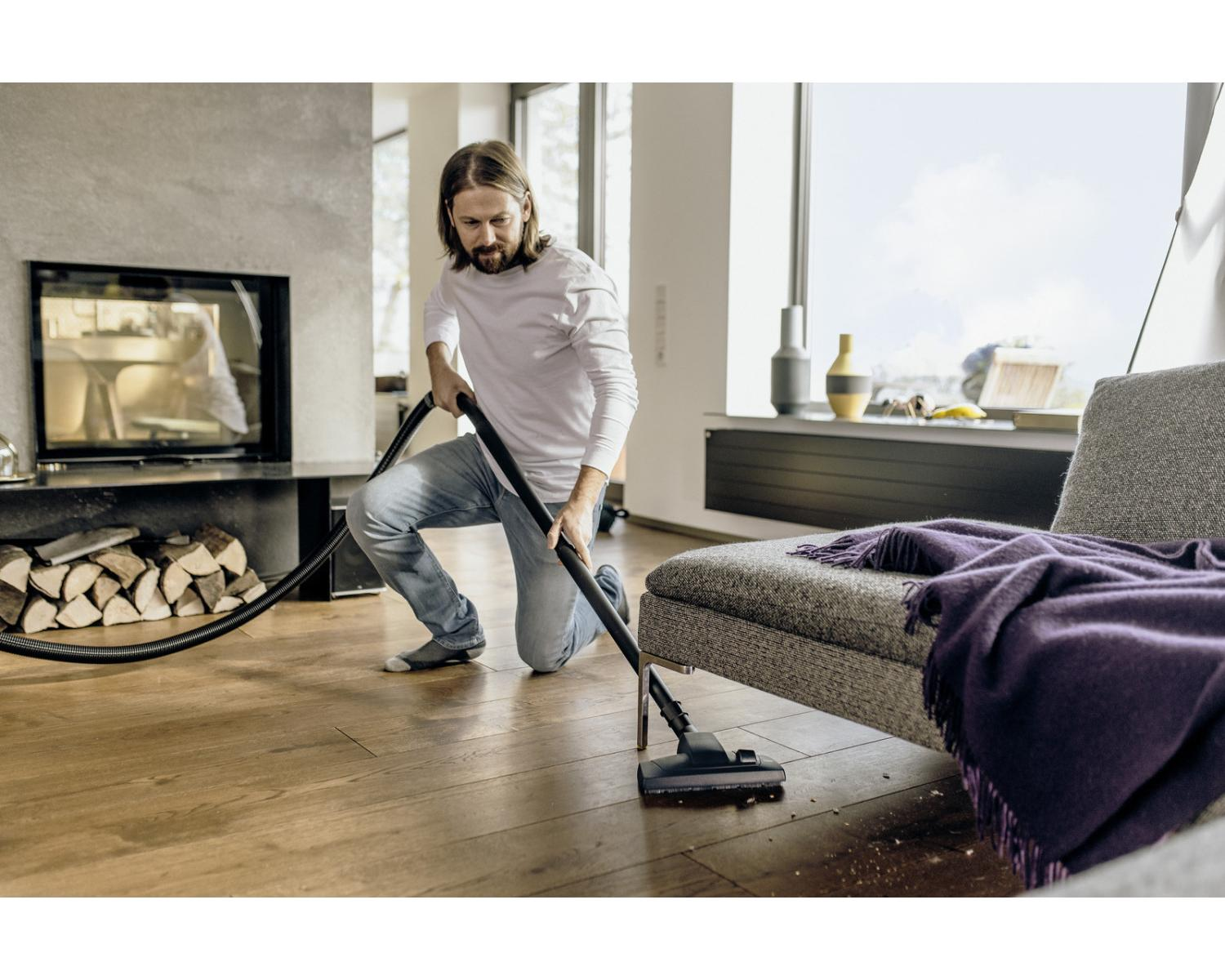 Hombre aspirando suelo de madera junto a chimenea y sofá con manta violeta.