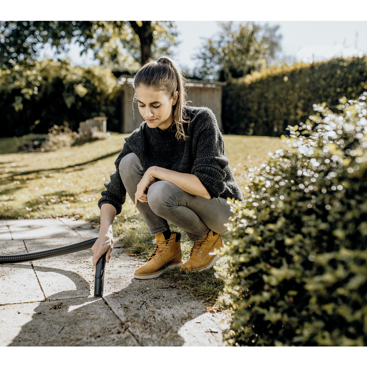 Donna pulisce sentiero con tubo in giardino. Outfit grigio, stivali gialli.