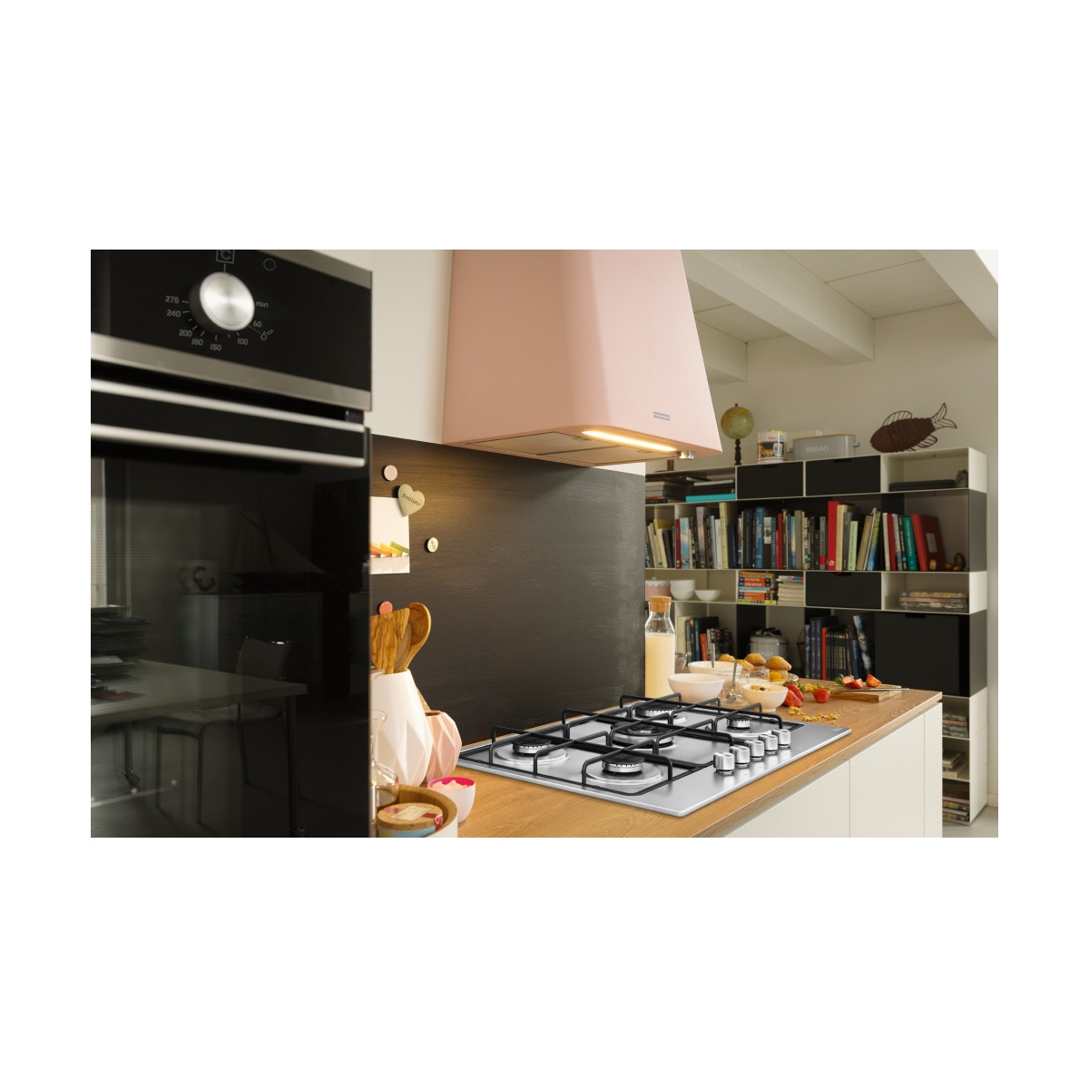 Una cucina moderna con piano cottura a gas, forno e cappa rosa. Scaffali con libri sullo sfondo.