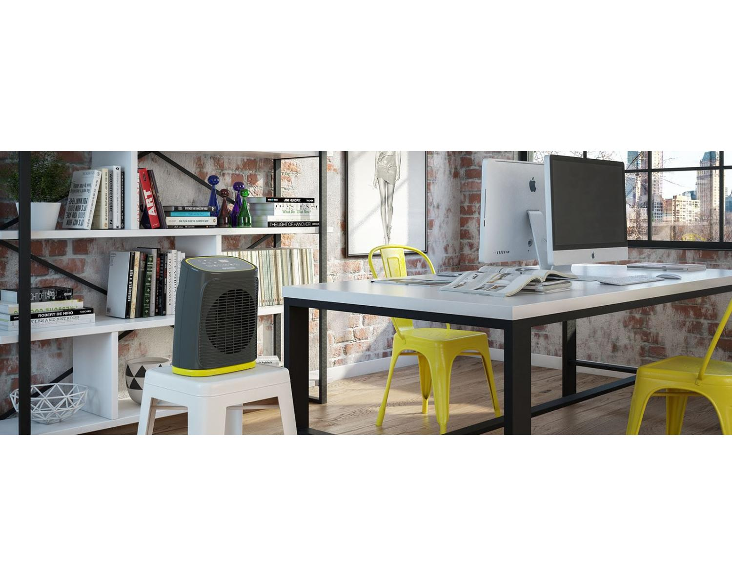 Ein Heizlüfter steht auf einem weißen Hocker neben einem Bücherregal. Ein Schreibtisch mit Computern und Stühlen ist im Hintergrund.