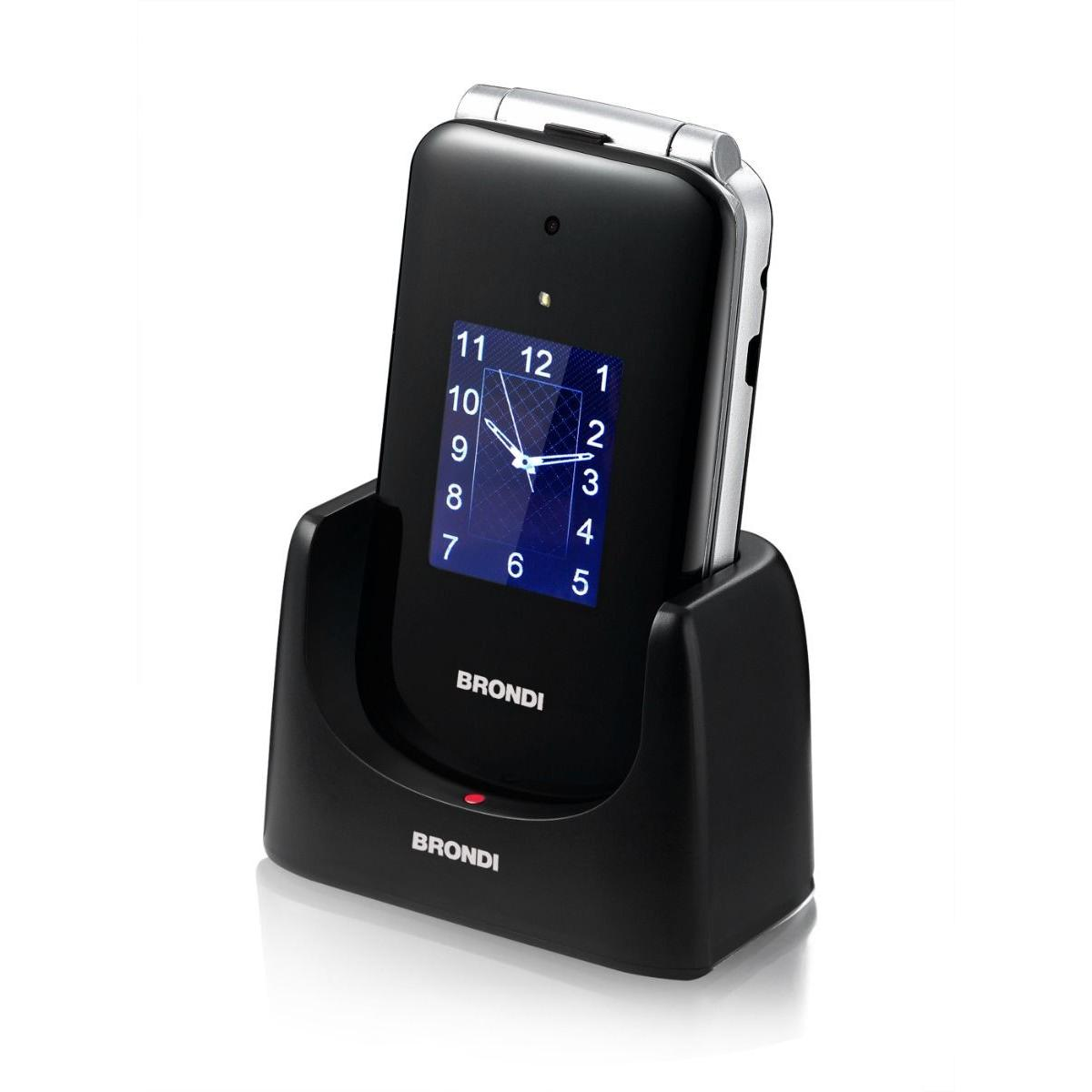 Un teléfono plegable negro Brondi está en su base de carga, mostrando una esfera de reloj.