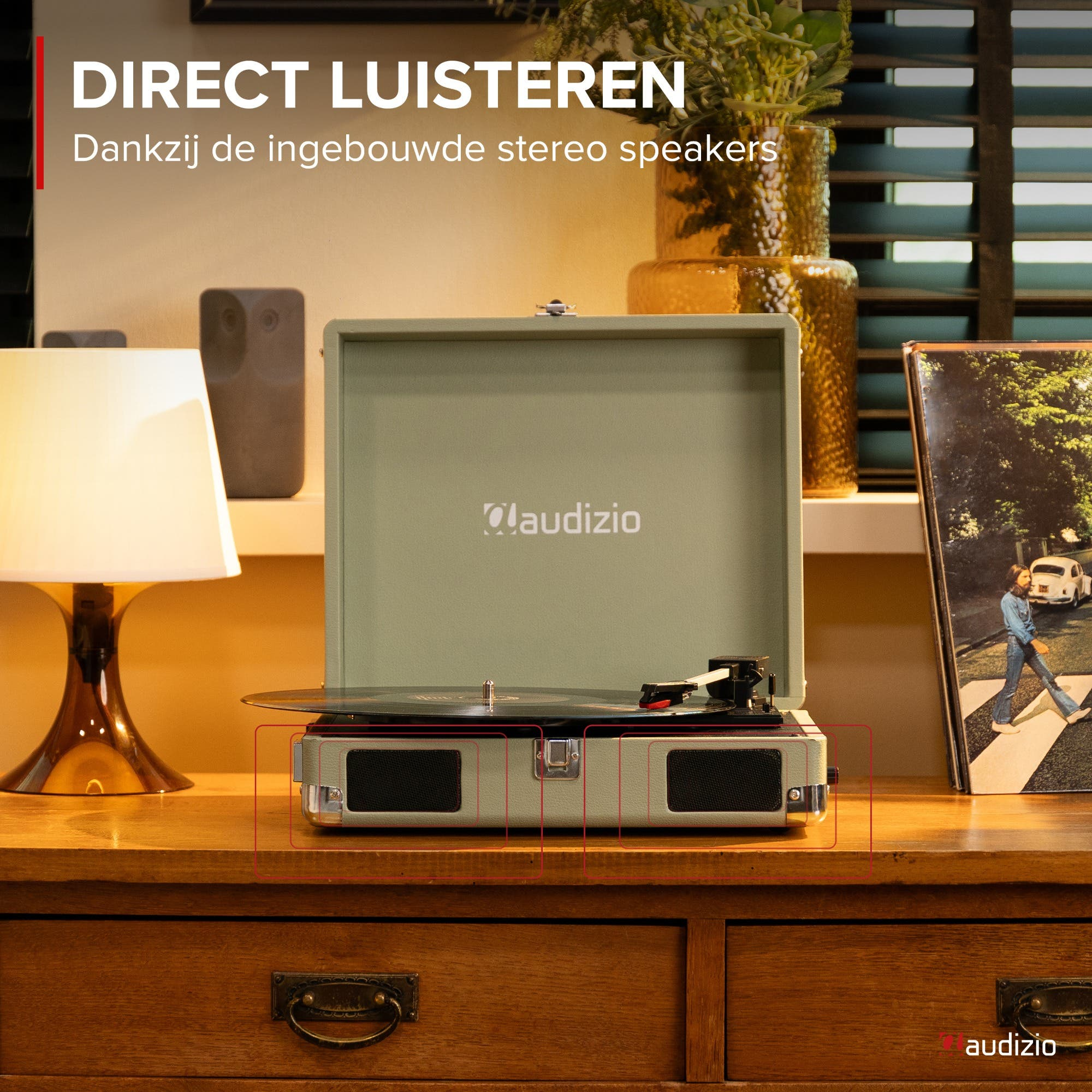 A record player with built-in speakers sits on a wooden desk with a lamp.