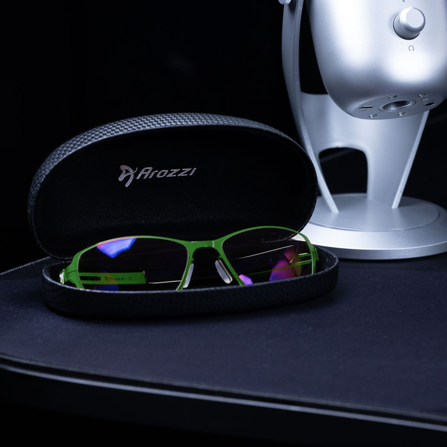 Grüne Brille in schwarzem Etui neben silbernem Mikrofon auf dunklem Untergrund.