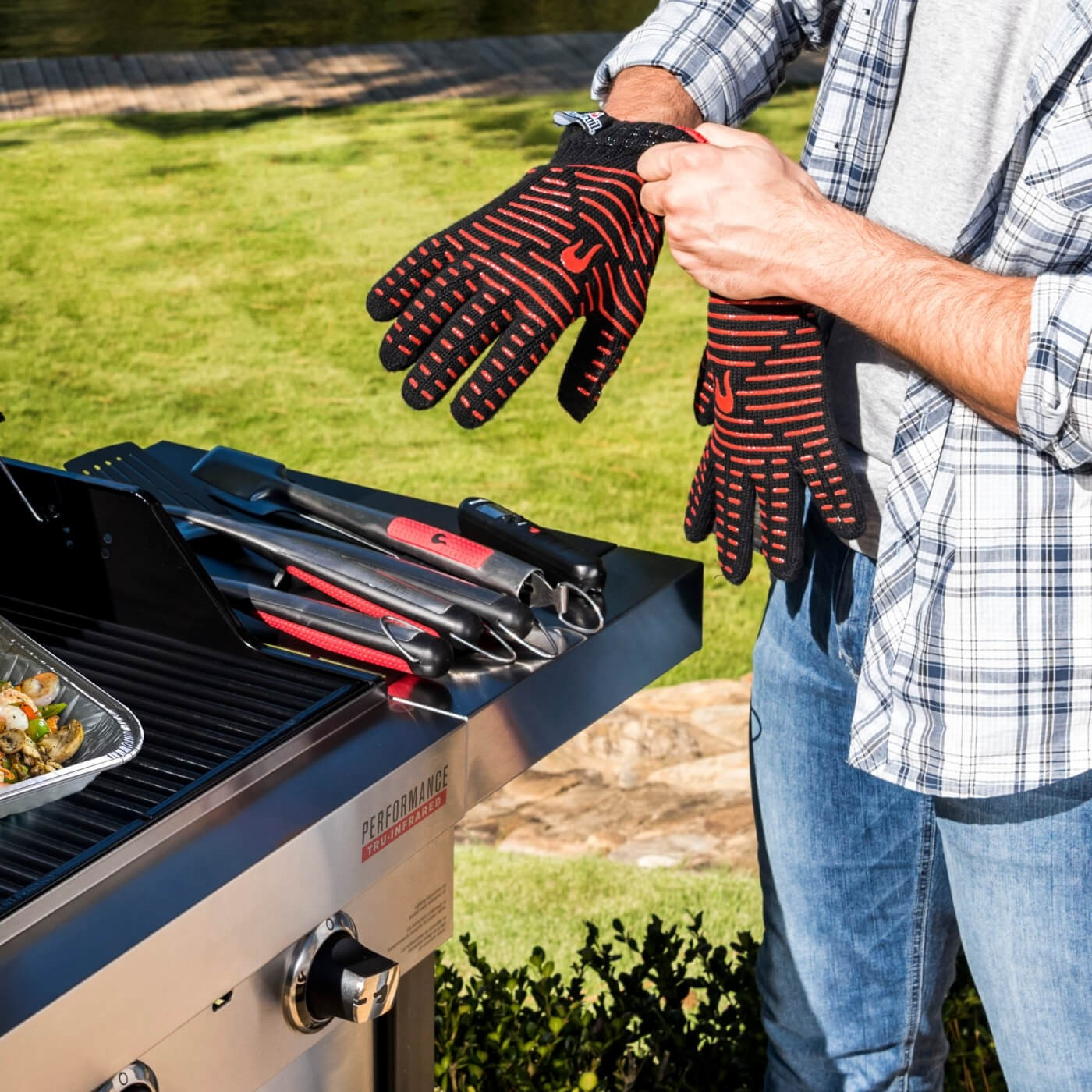Mann zieht Grillhandschuhe an, neben einem Grill mit Werkzeugen und Essen.