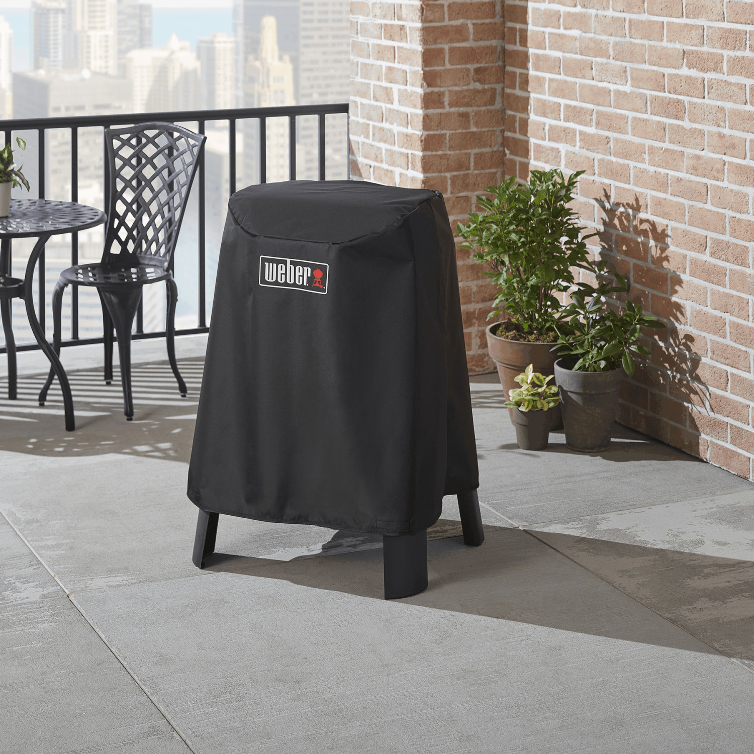 Funda negra para parrilla Weber en un balcón con vista a edificios de la ciudad.