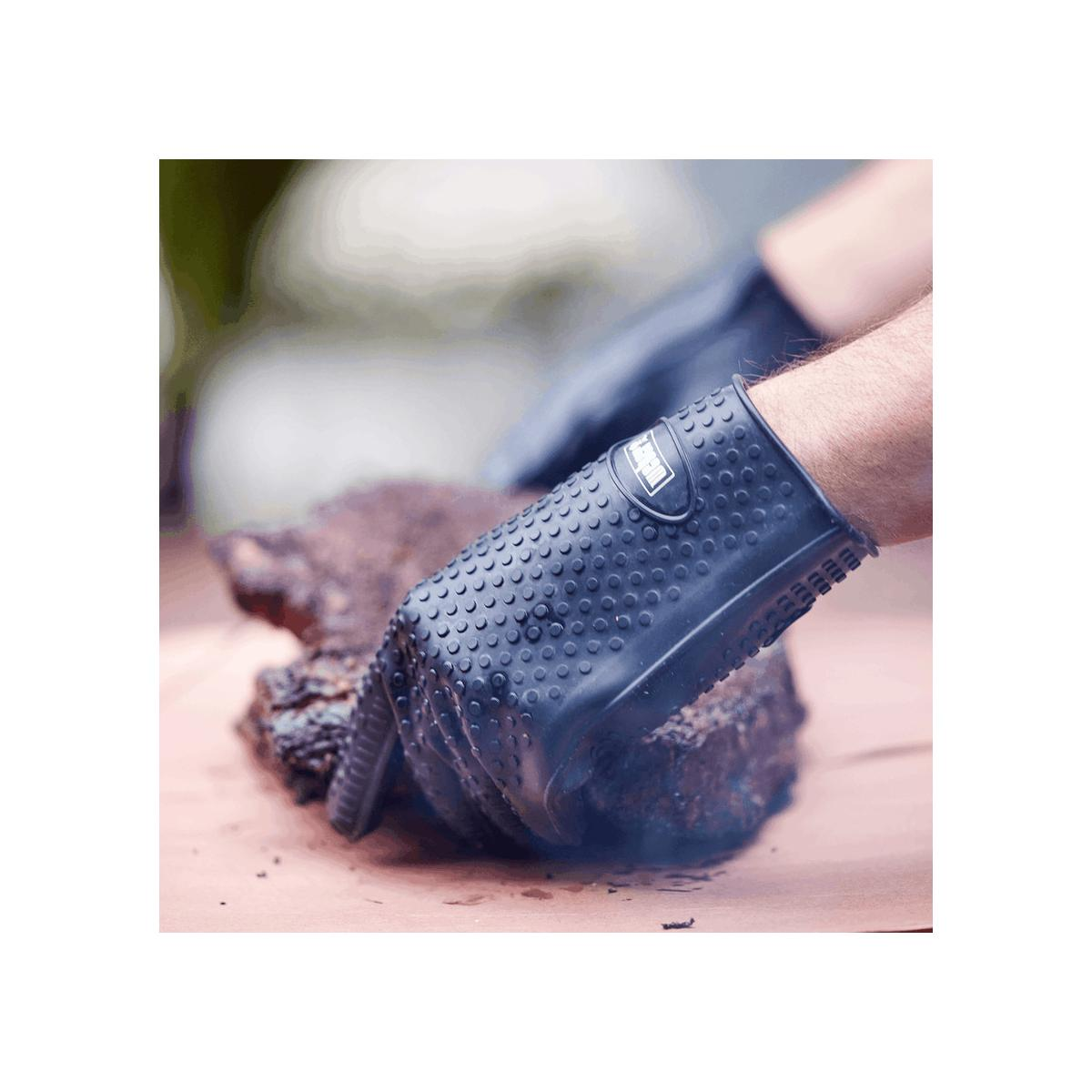 Eine Hand in einem schwarzen Grillhandschuh fasst ein dunkles Stück Fleisch an.
