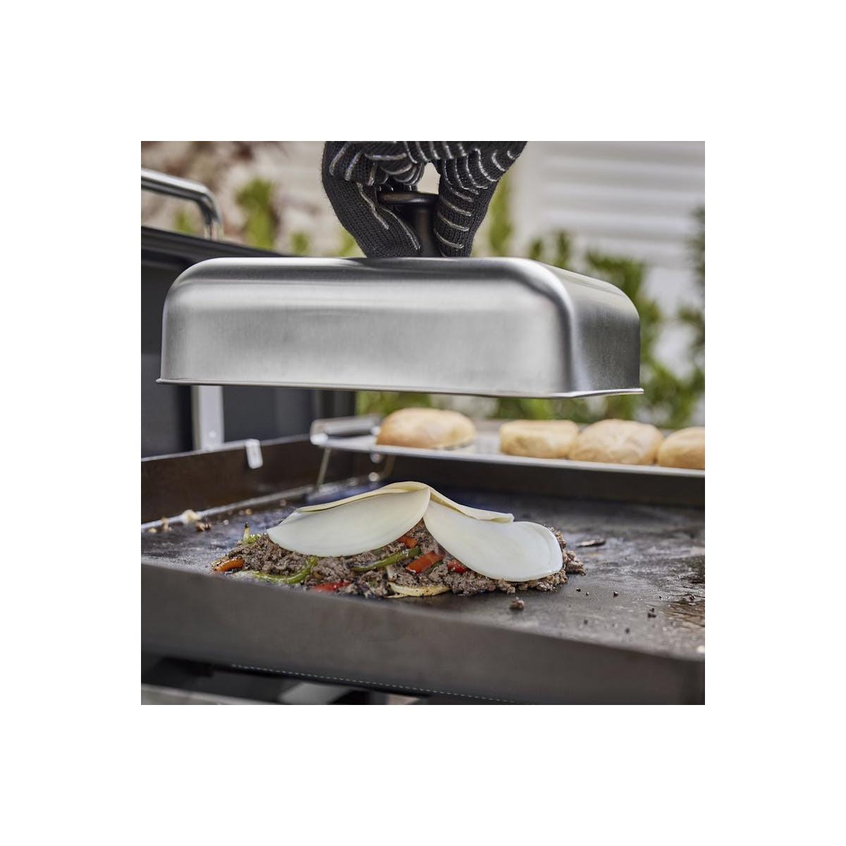 Ein Handschuh hält einen Metall-Dampfdom über einem Griddle mit Hackfleisch, Gemüse und Käse.
