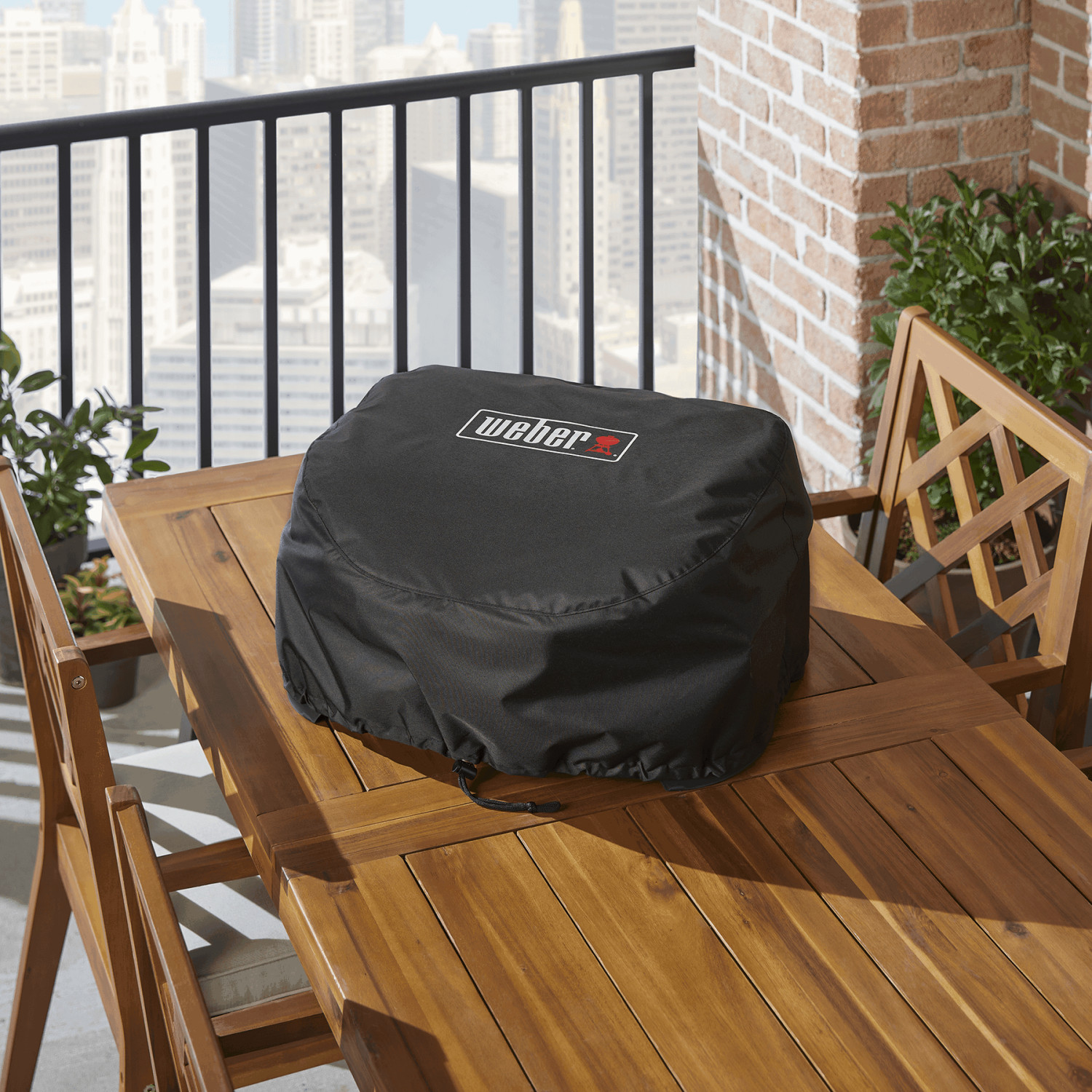 Funda negra Weber en mesa de madera con sillas, balcón y vista a la ciudad.