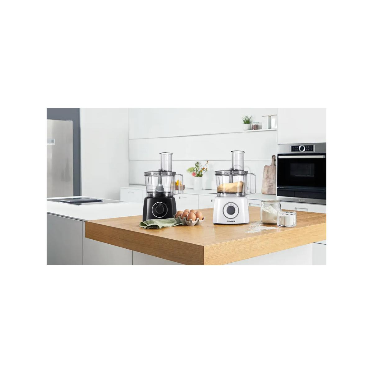 Due robot da cucina Bosch, uno nero e uno bianco, su un bancone di legno con uova e farina.