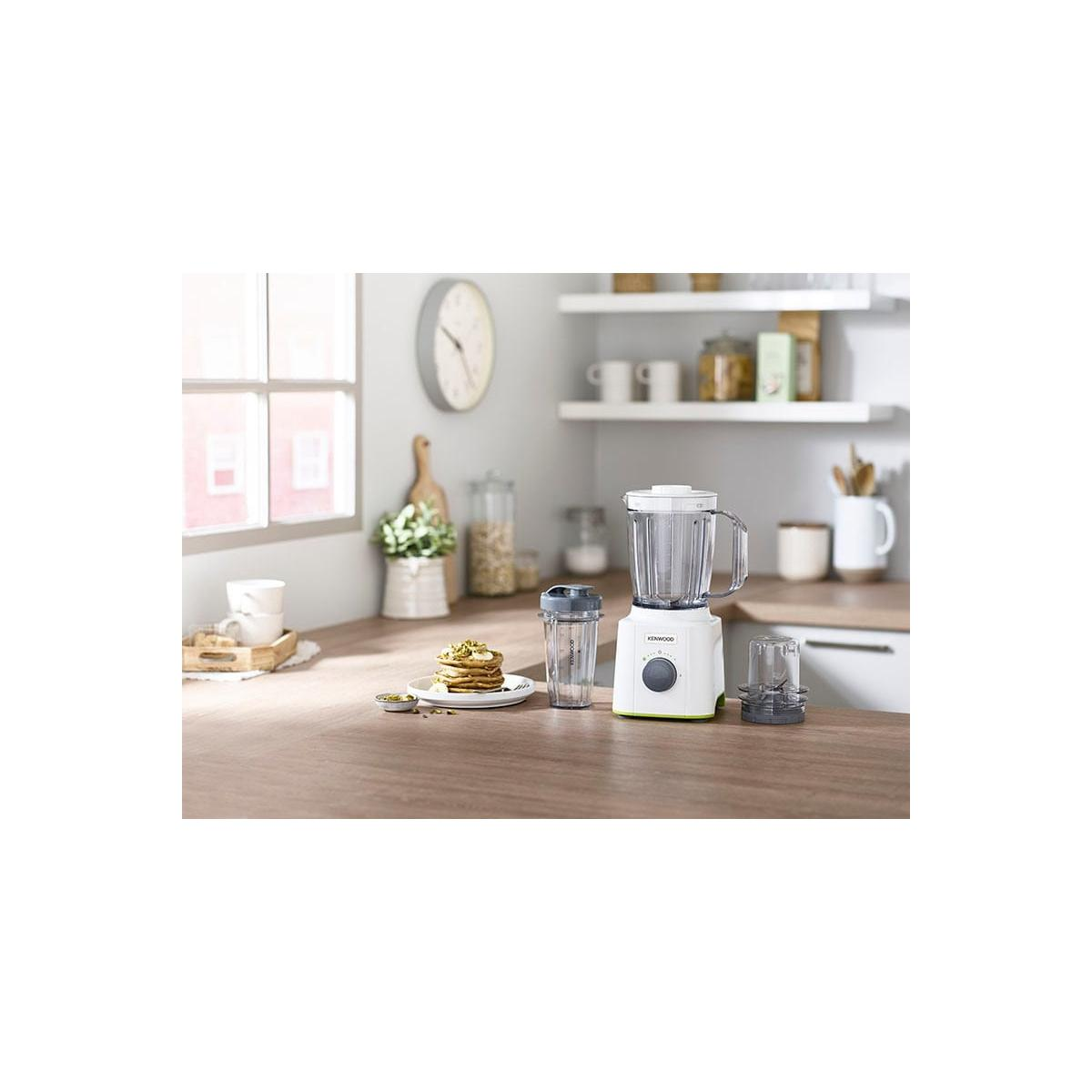 Een witte Kenwood blender met accessoires op een houten aanrechtblad. Een stapel pannenkoeken ernaast.