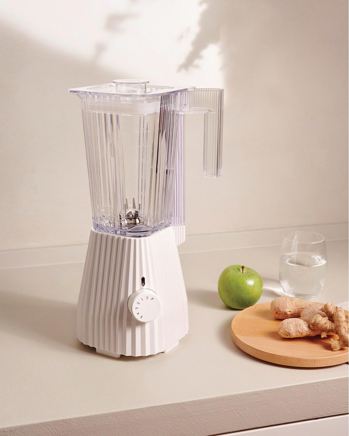 Un frullatore bianco con caraffa in vetro è su un piano di lavoro vicino a zenzero, mela e acqua.