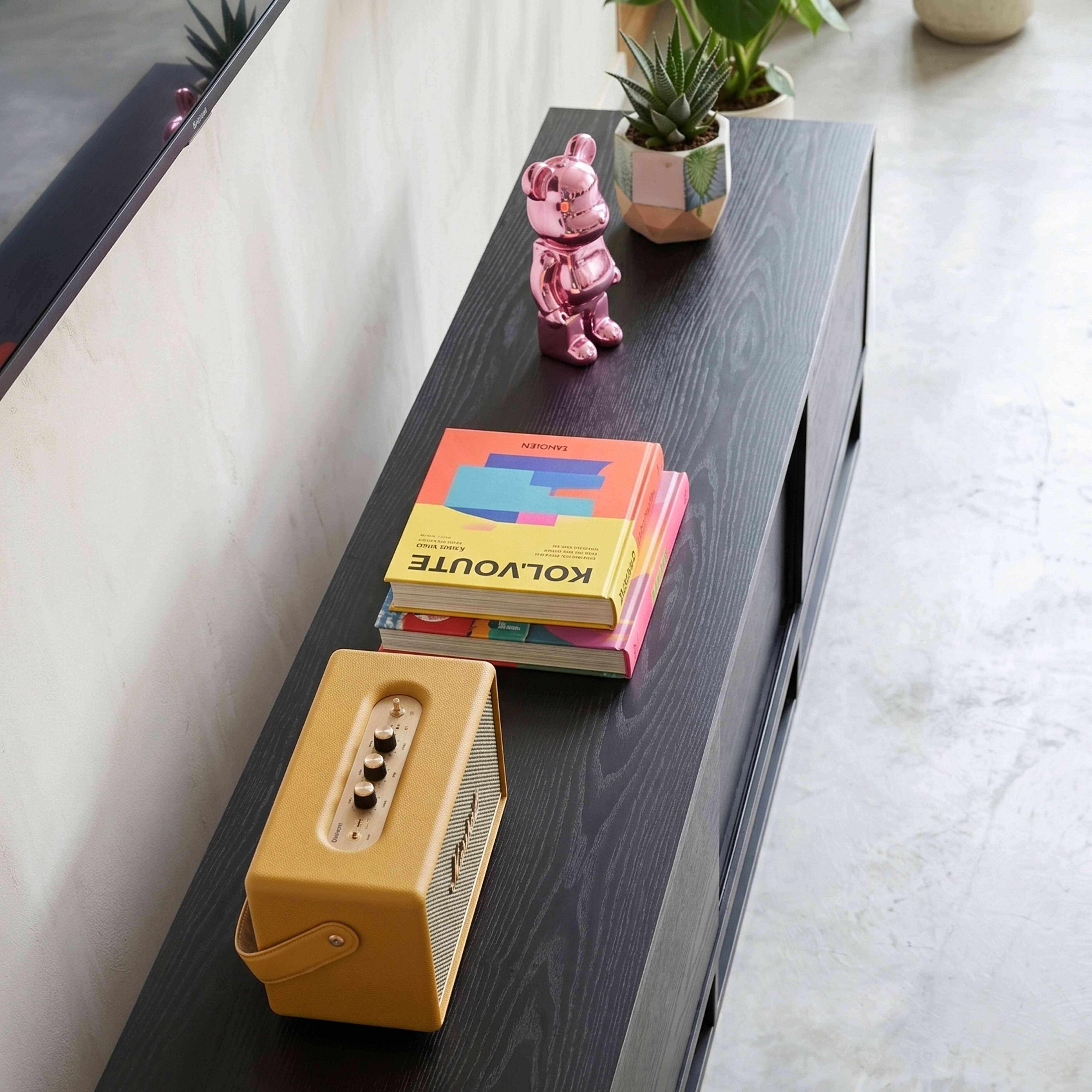 Ein rosa Teddybär, Bücher und ein gelber Lautsprecher auf einem dunklen Holzmöbel.