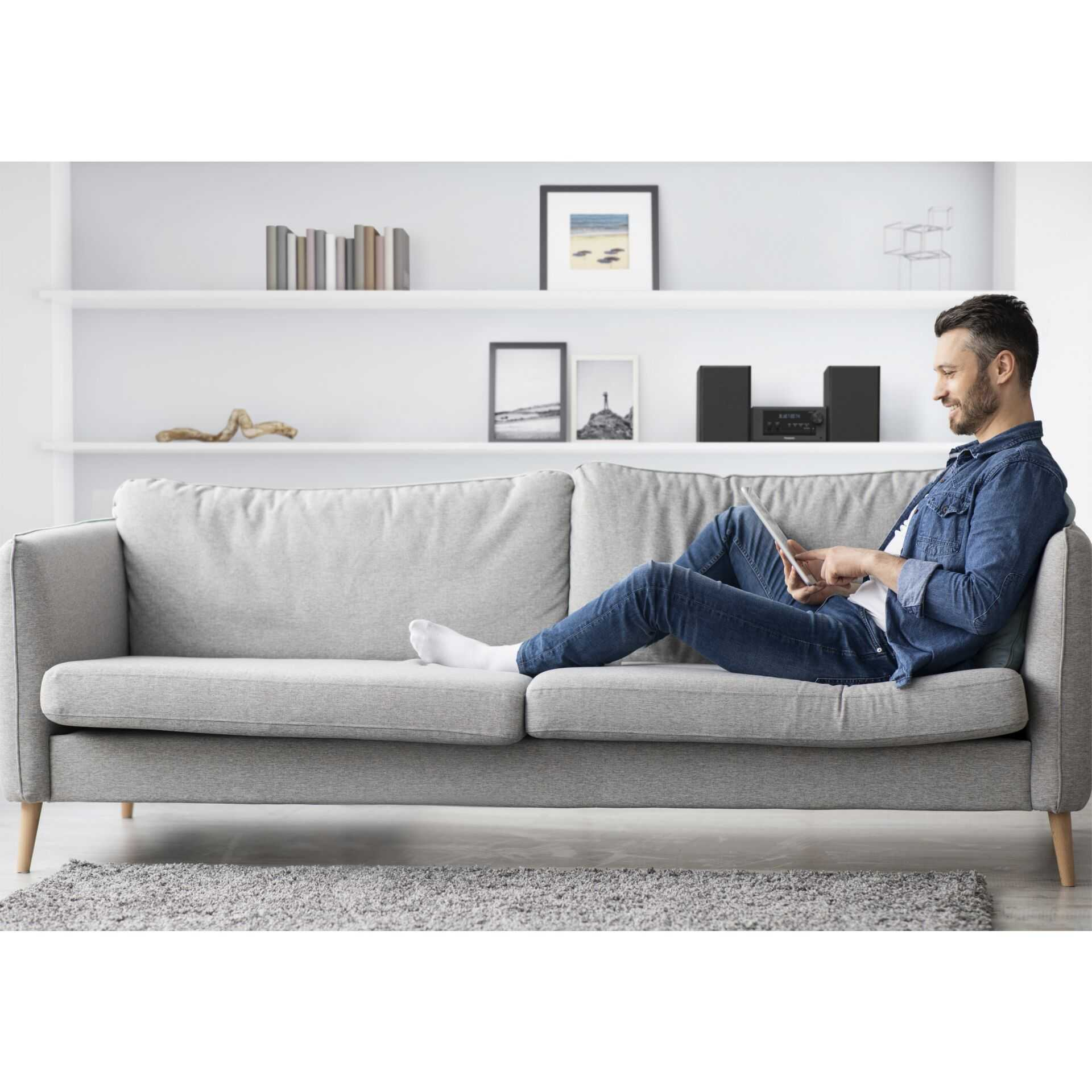 Ein Mann sitzt auf einem grauen Sofa und benutzt ein Tablet. Er trägt eine blaue Jeansjacke.
