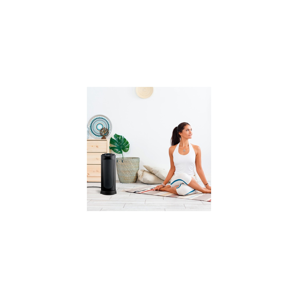 Een vrouw in witte sportkleding zit met gekruiste benen op een yogamat naast een zwarte torenverwarming.