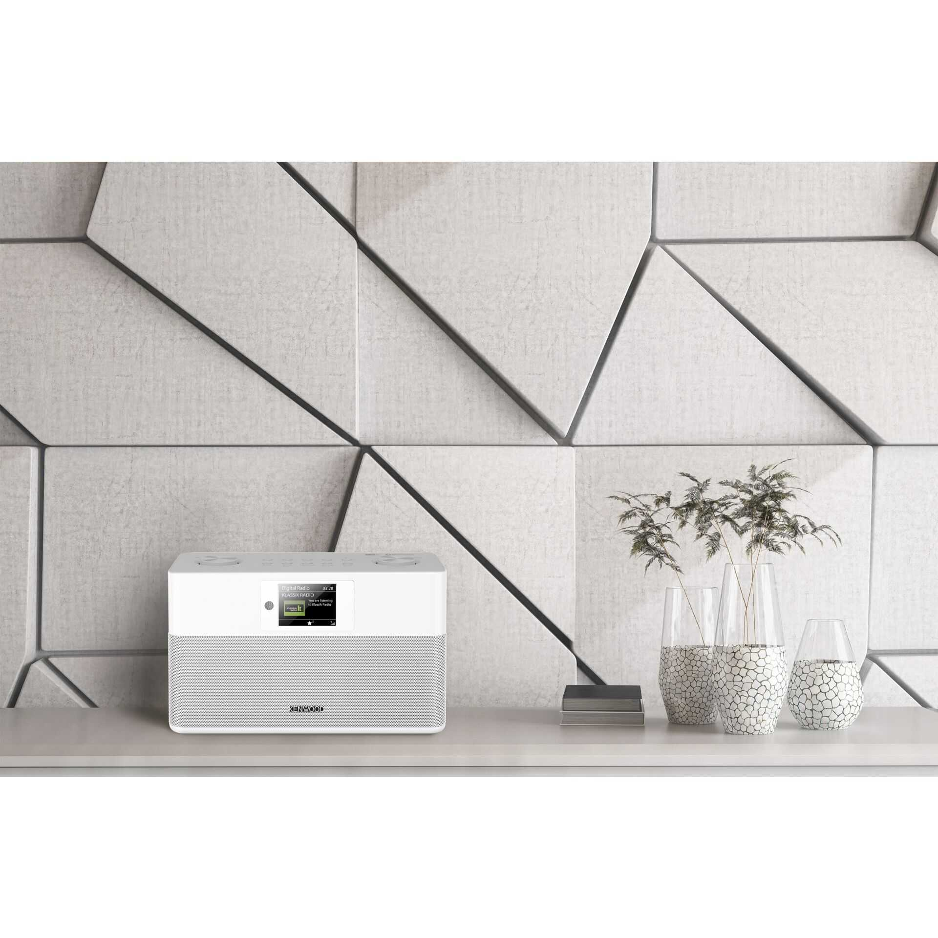Ein weißer digitaler Radio mit Bildschirm steht auf einem Regal vor einer geometrischen Wand.