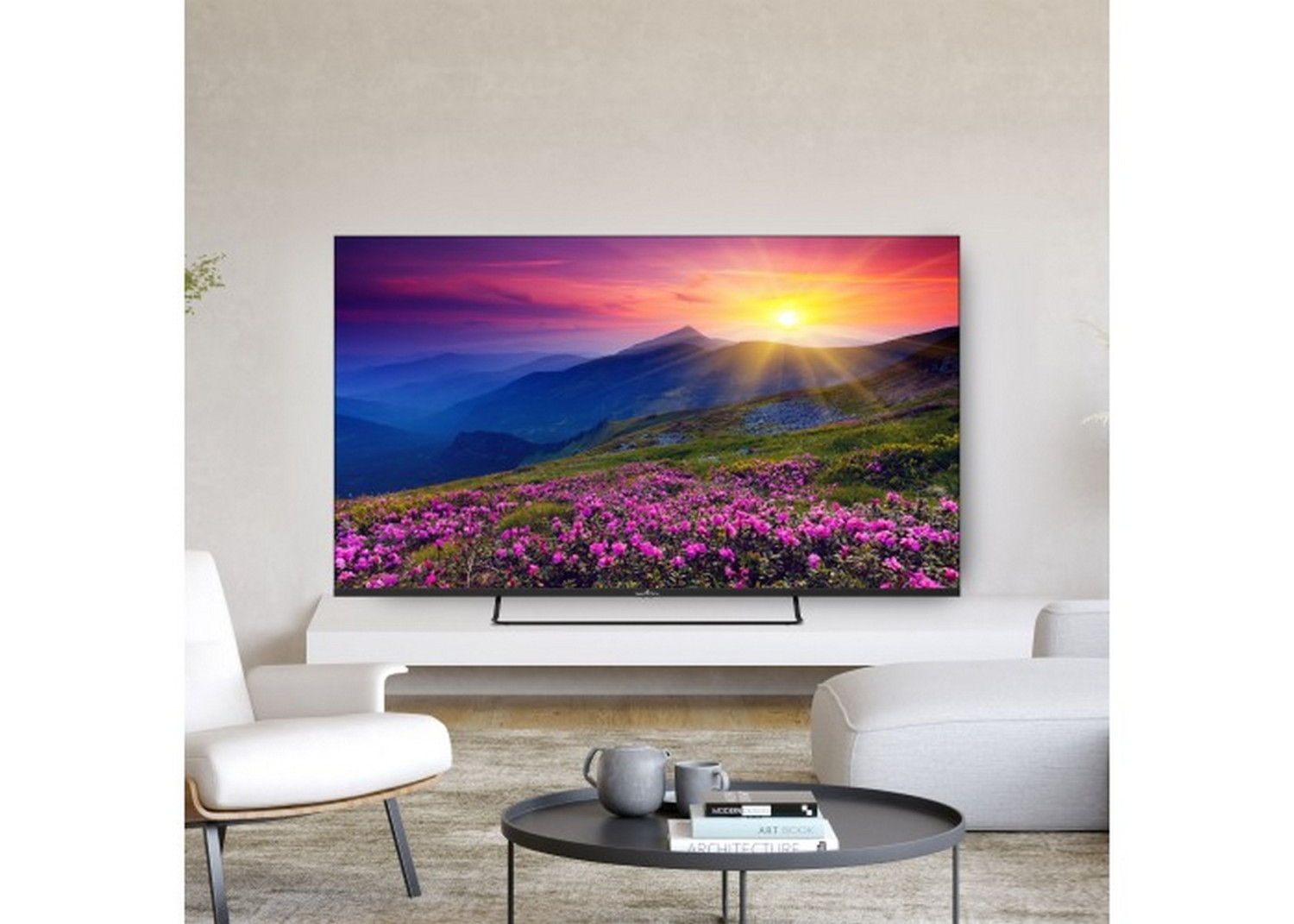 Una TV mostra un paesaggio montano con fiori viola al tramonto. Sedie bianche e tavolino nelle vicinanze.