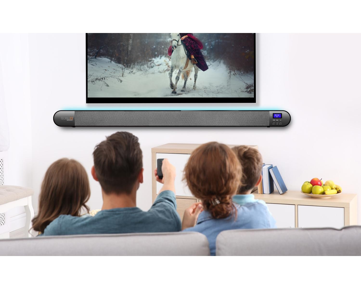 Famiglia guarda TV con soundbar, persona tiene telecomando, cavallo sullo schermo.