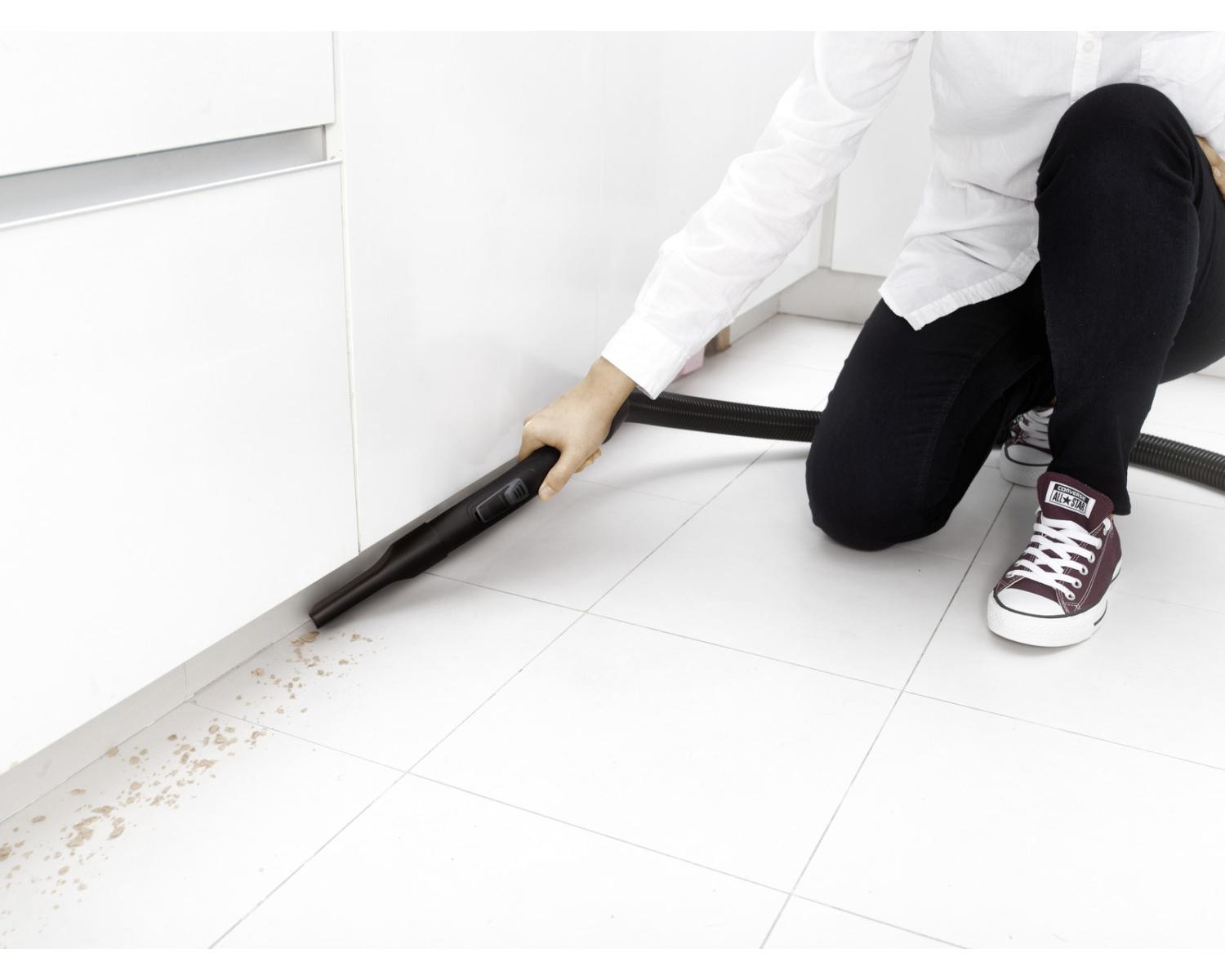 Eine Person saugt Schmutz vom weißen Fliesenboden mit einem schwarzen Handstaubsauger.