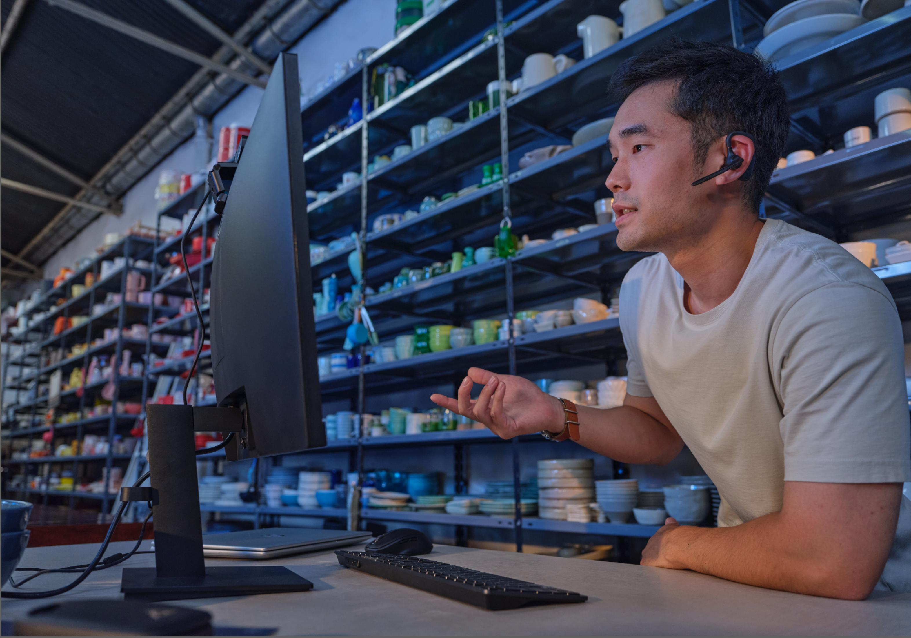 Man met headset praat bij computer, schappen met aardewerk op achtergrond.