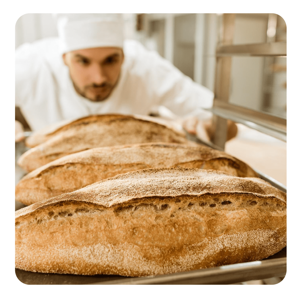 Ein Bäcker in weißer Kleidung prüft frisch gebackenes Brot auf einem Blech.