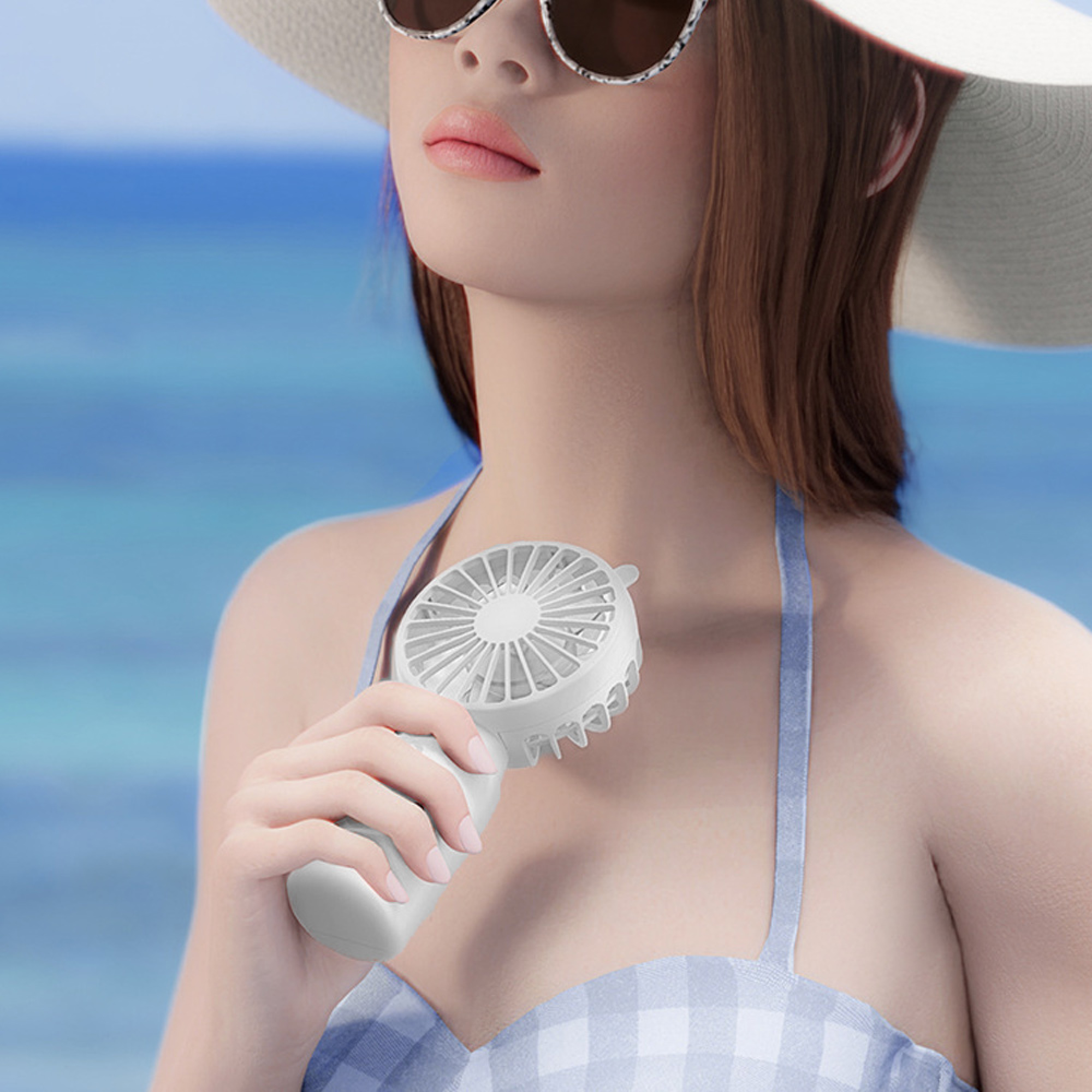 Frau mit Sonnenbrille und Hut hält einen weißen Ventilator am Strand.