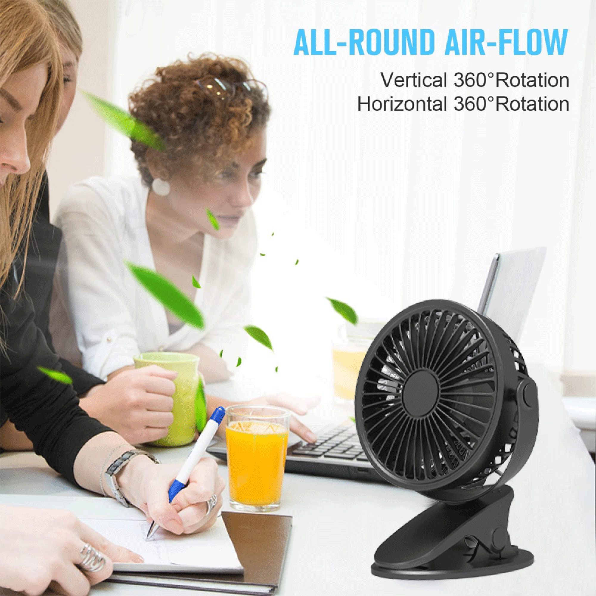 Ein schwarzer Tischventilator steht neben einem Laptop, zwei Frauen arbeiten.
