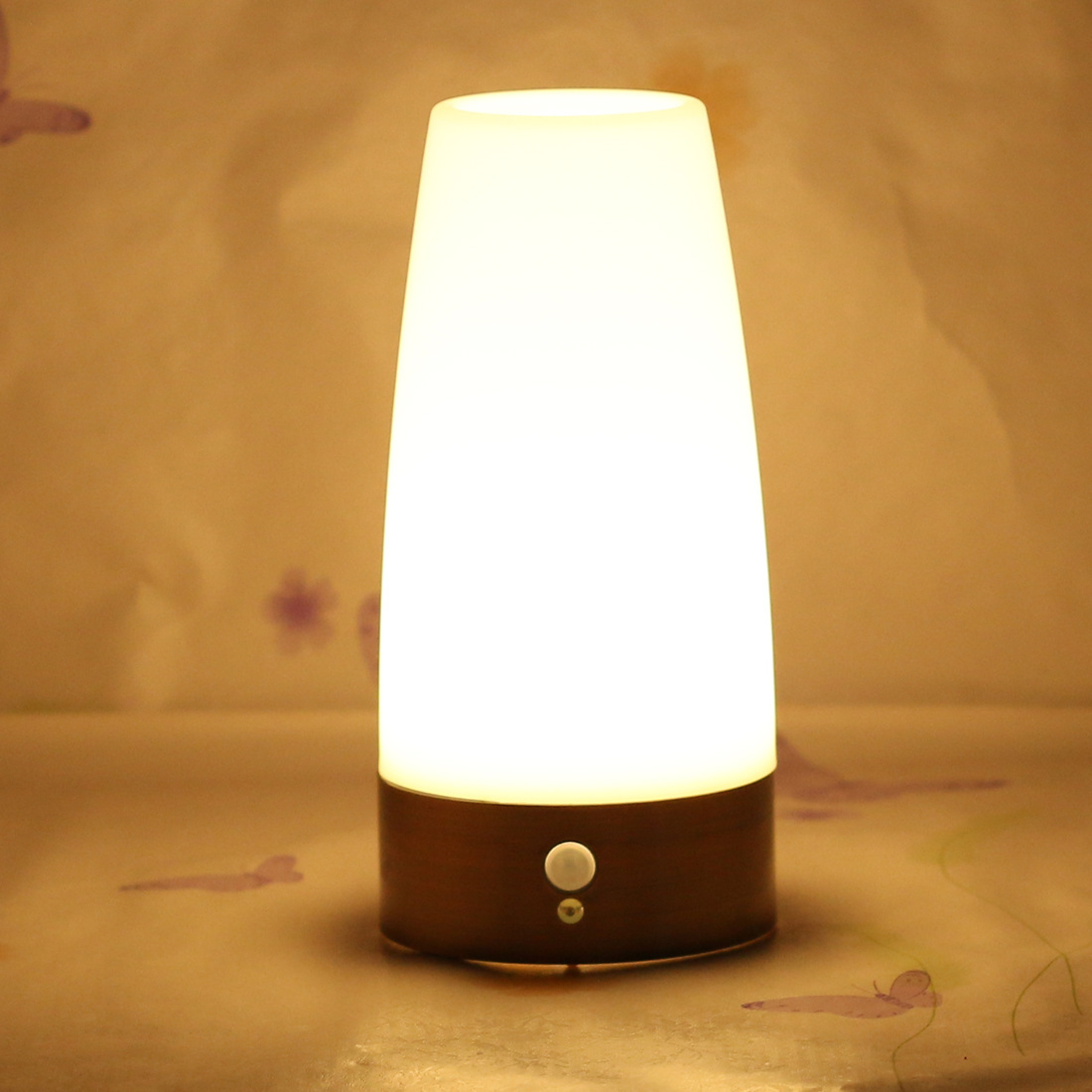 Eine Nachttischlampe mit einem braunen Sockel und einem weißen Leuchtschirm. Hinten ist eine gemusterte Wand.