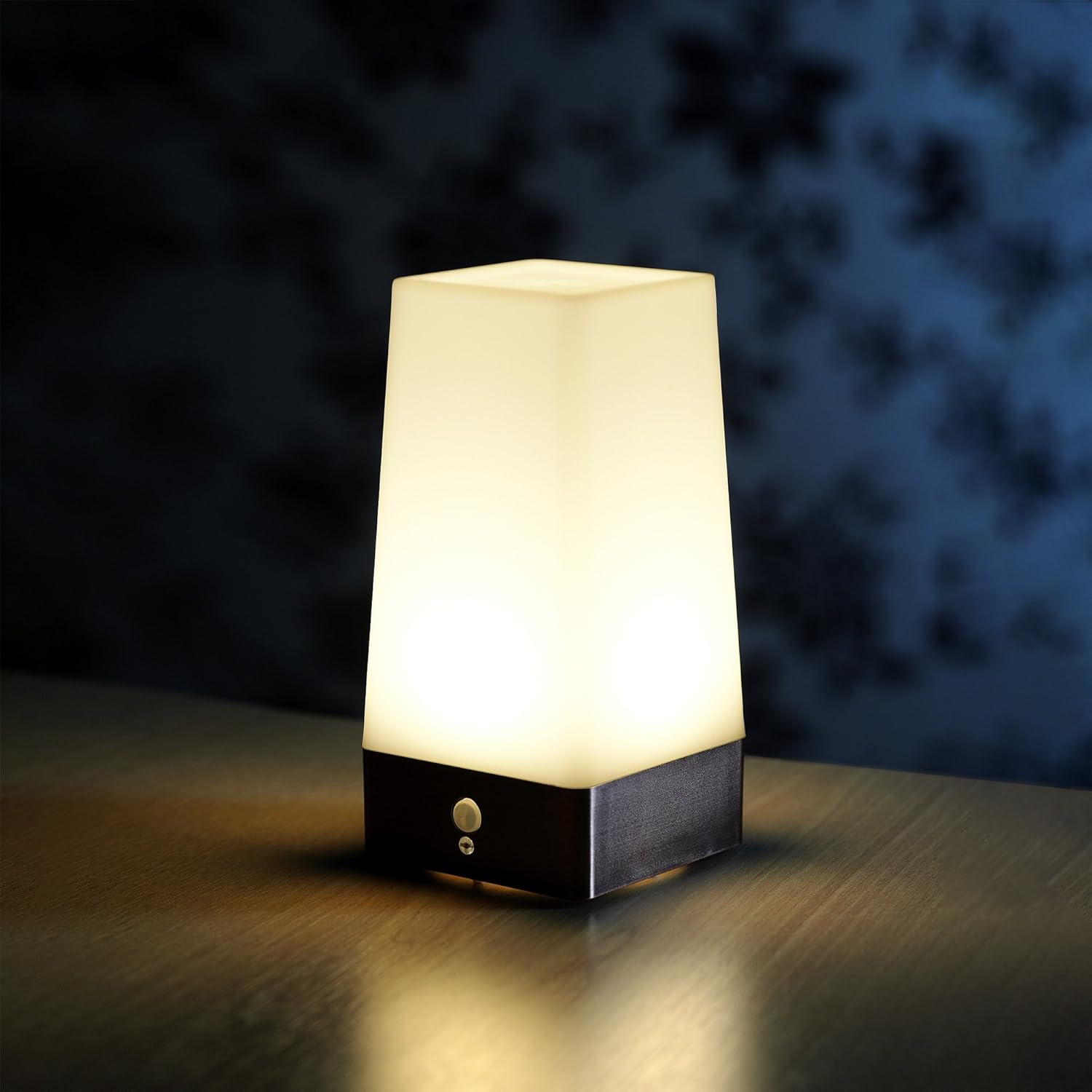 Eine beleuchtete weiße Lampe mit dunklem Sockel steht auf einem Holztisch.